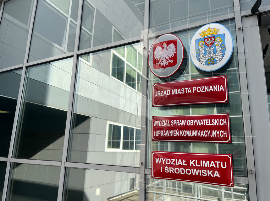 Ilustracja przedstawia wejście do urzędu. Na tablicach widoczne godło państwowe, herb miasta, a także nazwy - Urząd Miasta Poznania, Wydział Spraw Obywatelskich i Uprawnień Komunikacyjnych oraz Wydział Klimatu i Środowiska.