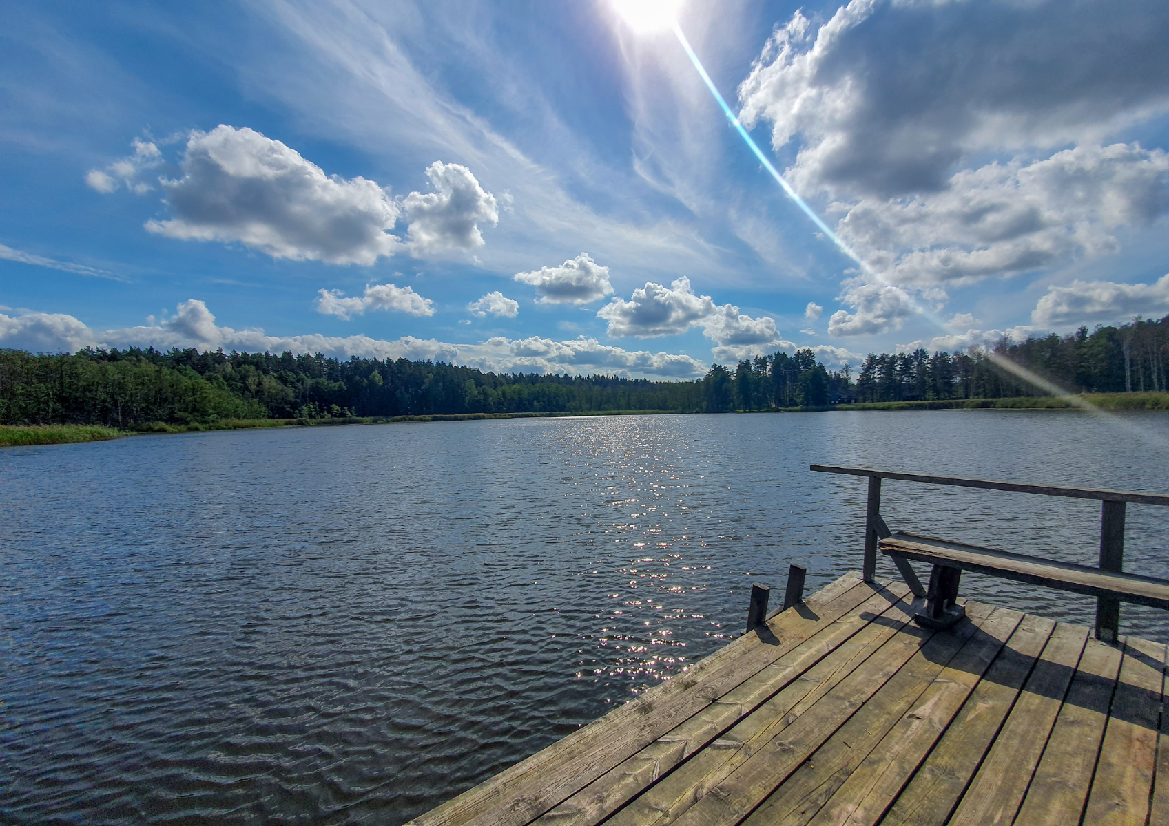 Jezioro z kawałkiem pomostu, w tle znajduje się brzeg z drzewami. Pogoda jest słoneczna, woda niebieska.
