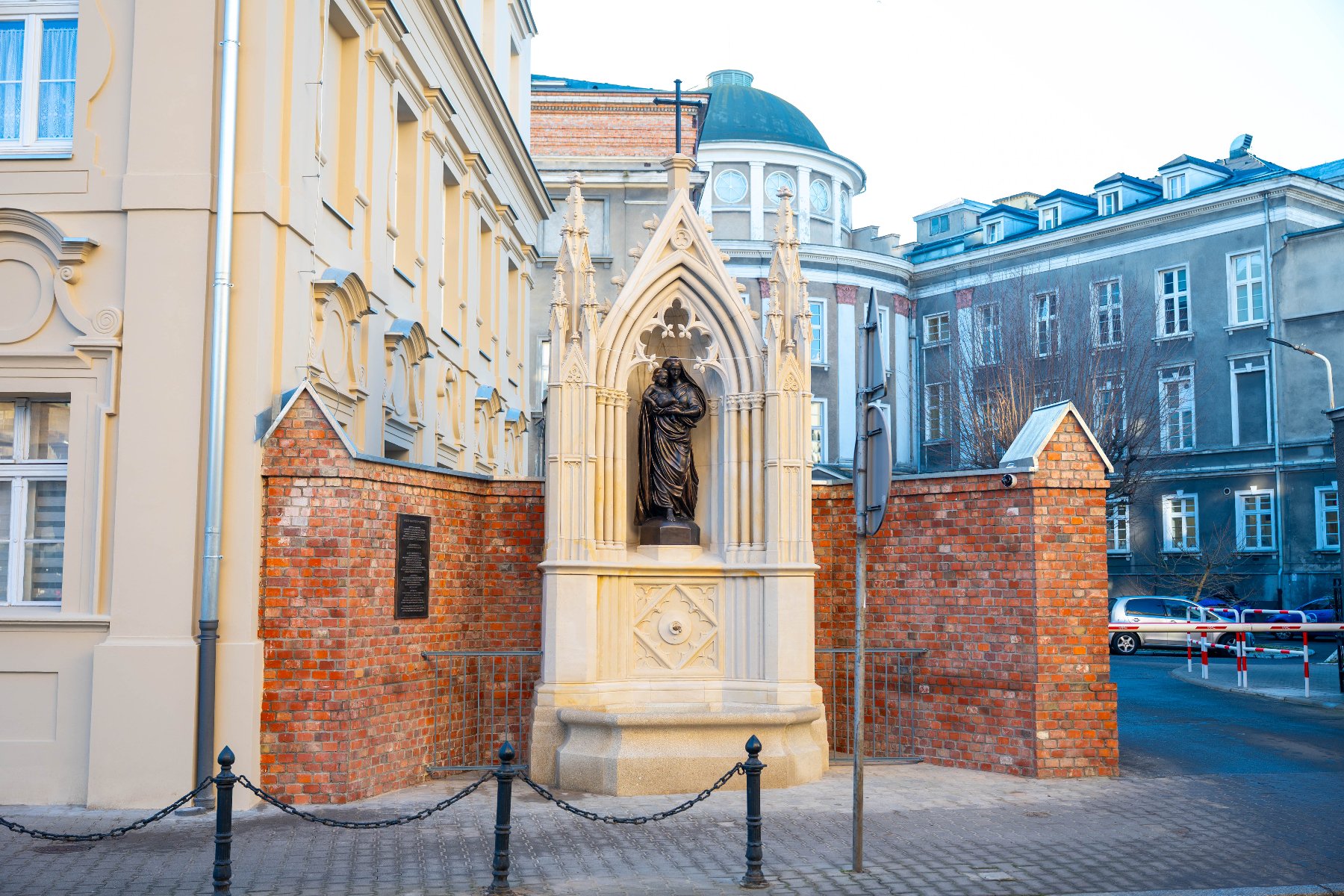 Kamienna figura Matki Bożej z Dzieciątkiem umieszczona w neogotyckiej kapliczce wbudowanej w ceglany mur. Po lewej stronie jasna elewacja budynku, w tle widoczna kopuła gmachu biblioteki oraz inne budynki miejskie. Na pierwszym planie fragment chodnika i niski metalowy płotek z łańcuchem.