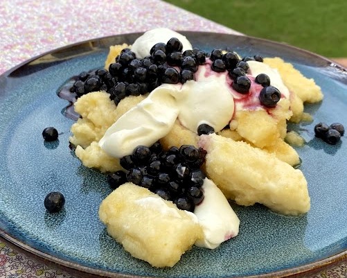 Tradycyjne leniewe z dodatkiem jagód, fot. AL Kluski leniwe z jagodami i kleksem śmietany na niebieskim talerzu.