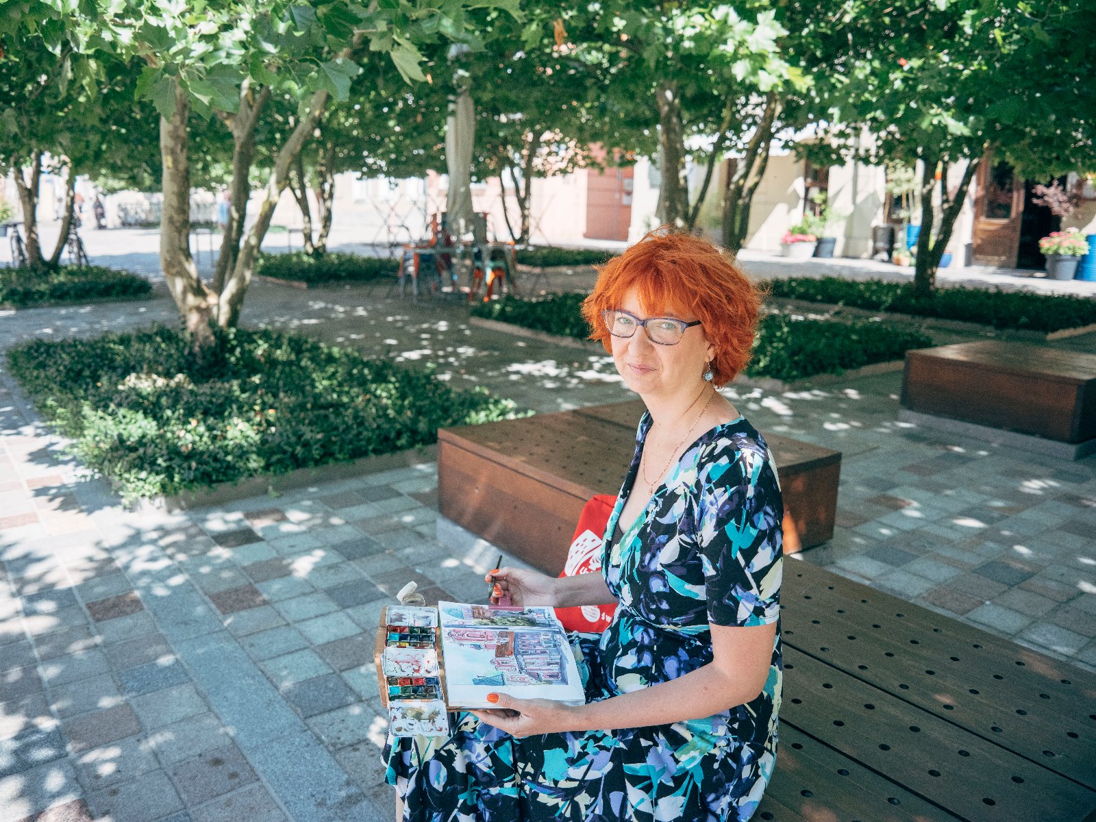 Kobieta siedzi na ławce, na miejskim placu wśród zieleni i maluje.