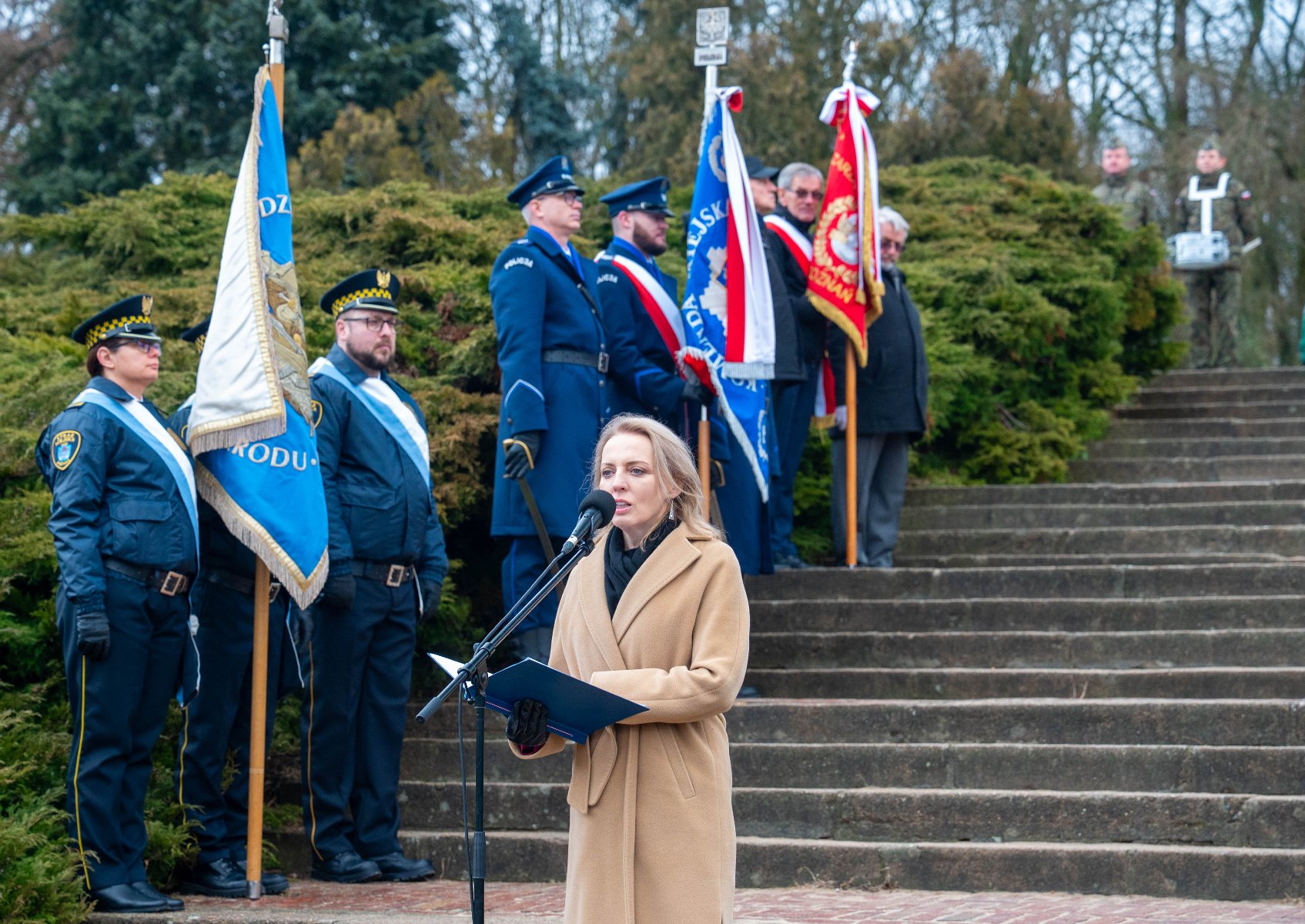 Kobieta w beżowym płaszczu przemawia do mikrofonu, trzymając w dłoniach otwarty niebieski folder. Stoi na tle kamiennych schodów, na których ustawione są poczty sztandarowe. Po lewej stronie widoczni są funkcjonariusze Straży Miejskiej w ciemnych mundurach z błękitnymi szarfami, a w głębi policjanci oraz mężczyzna w cywilnym ubraniu trzymający czerwony sztandar.