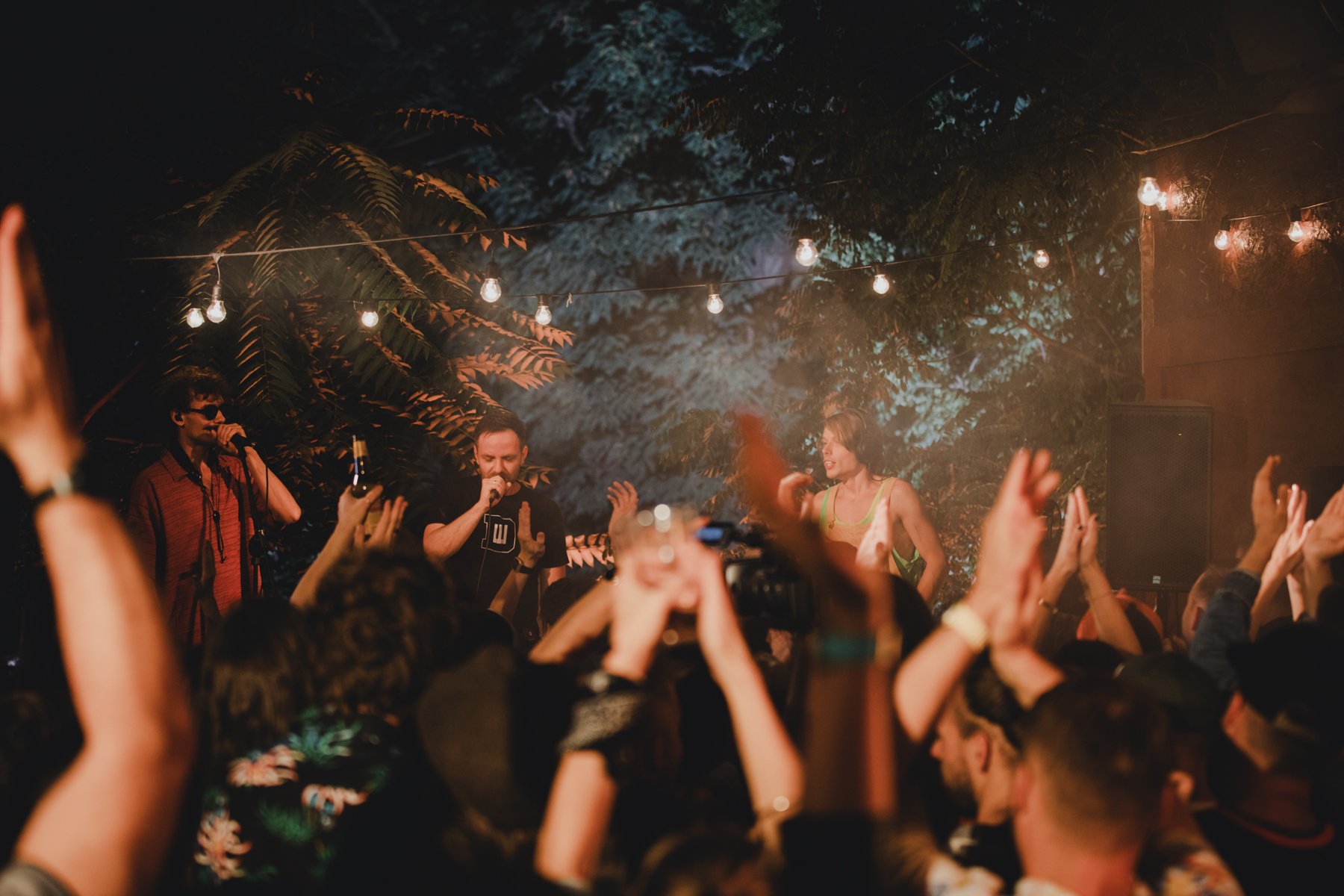 fot. Beata Wencławek - koncert TAXI w Barze u Gosi 2023 Koncert w pomieszczeniu oświetlonym wiszącymi żarówkami i czerwonym światłem. Na scenie trzech artystów śpiewa do mikrofonu. Na pierwszym planie widoczny tłum publiczności z uniesionymi rękami.