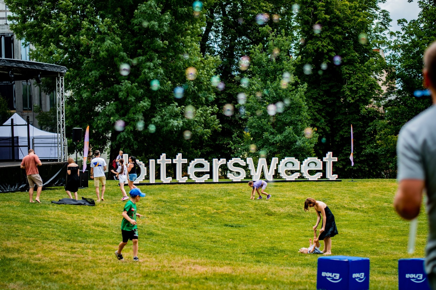 Od 14 do 16 sierpnia na terenie Cytadeli odbędzie się pierwsza edycja międzynarodowego BitterSweet Festival Ludzie bawią się na zielonej polanie. Na trawie znajdują się duże, białe litery układające się w napis "bittersweet". W tle widać scenę, drzewa i latające bańki mydlane.