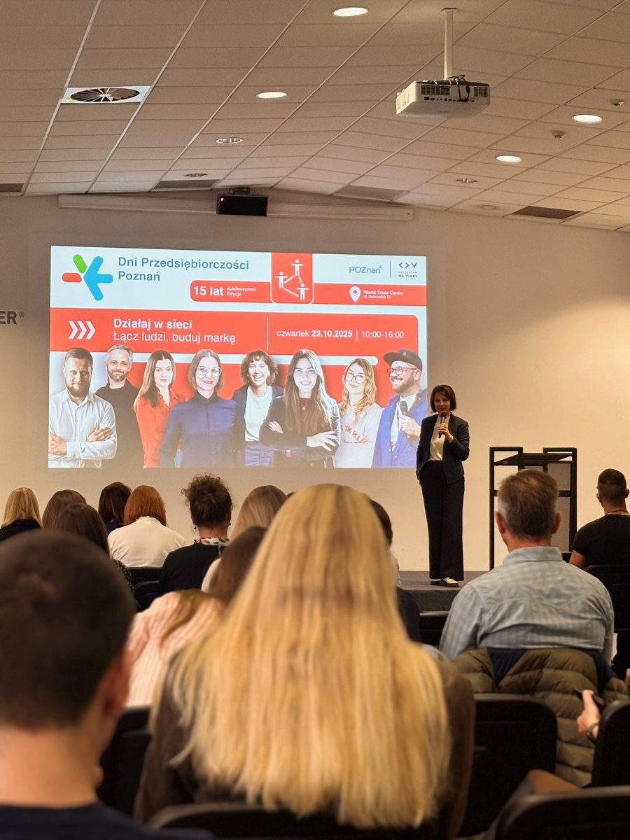 Mężczyzna- Kuba Łuka, ubrany w czarną kurtkę i z identyfikatorem konferencyjnym, stoi na tle ścianki Dni Przedsiębiorczości Poznań