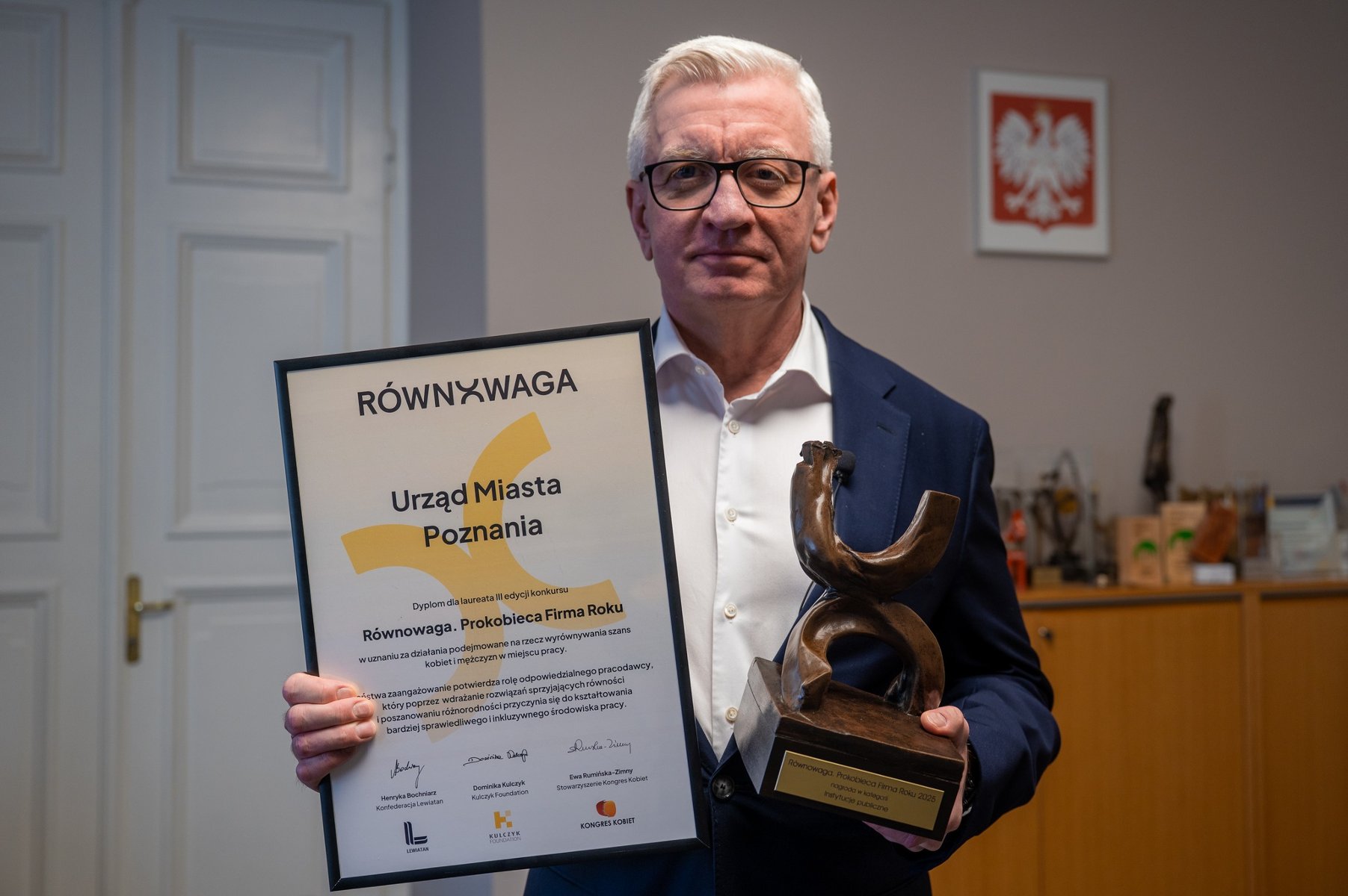 Mężczyzna w okularach i granatowej marynarce trzyma dyplom oraz brązową statuetkę. Na dyplomie napisy: "RÓWNOWAGA", "Urząd Miasta Poznania", "Równowaga. Prokobieca Firma Roku". W tle na ścianie godło Polski, po lewej białe drzwi.