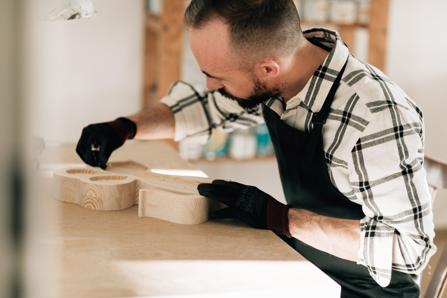 Mężczyzna z brodą, ubrany w koszulę w kratę i czarny fartuch ochronny, pracuje przy drewnianym stole warsztatowym. Ma założone czarne rękawice robocze i precyzyjnie obrabia dłońmi duży, jasny element wycięty z drewna o wyraźnym usłojeniu.