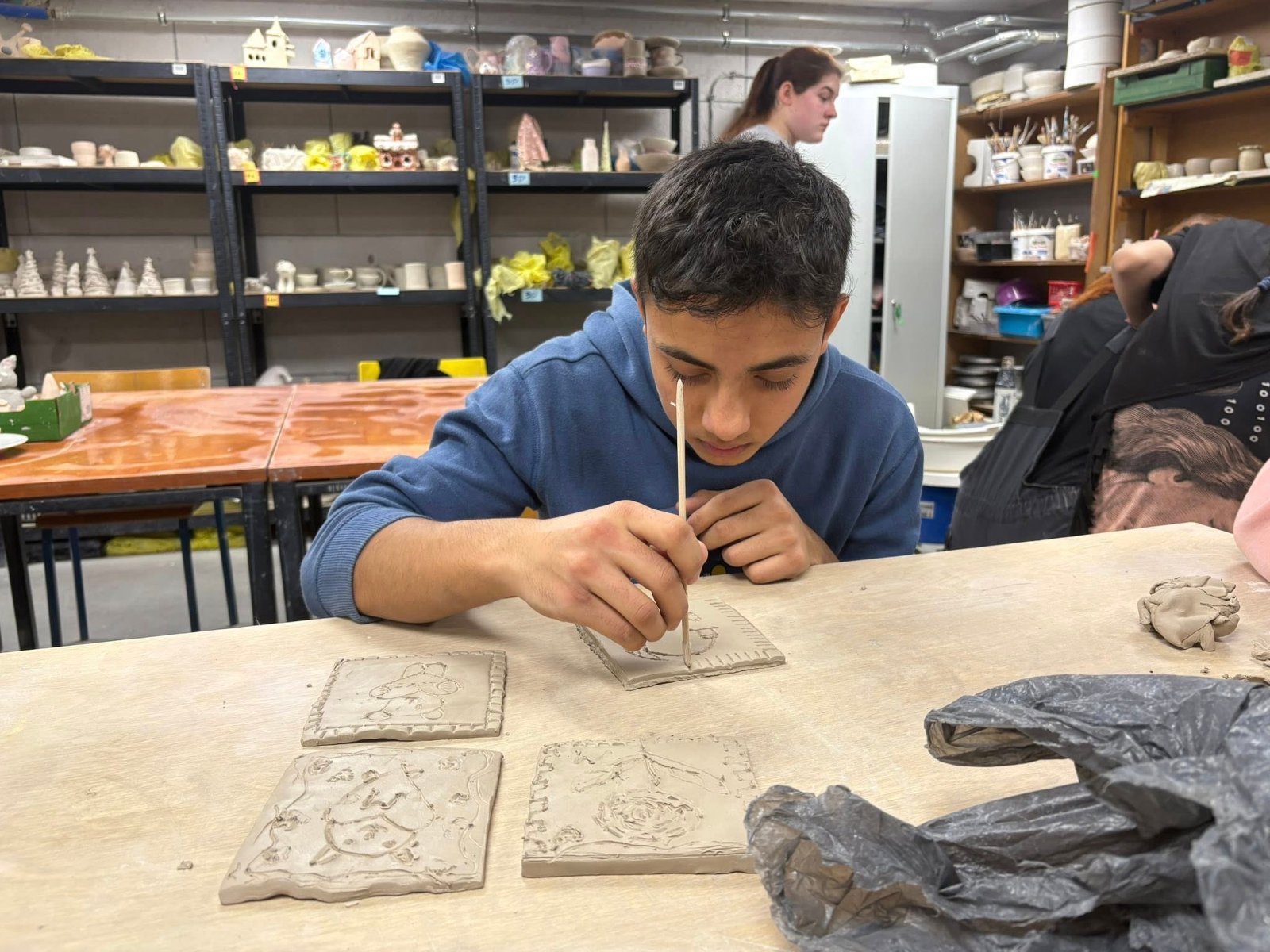 Młody mężczyzna pracuje nad glinianą płytką, formując ją rękami na stole roboczym.