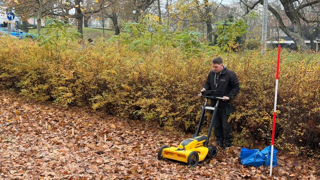 Młody mężczyzna w czarnej kurtce stoi w parku pełnym opadłych, brązowych liści. Obsługuje żółto-czarne urządzenie pomiarowe na kółkach (prawdopodobnie georadar lub skaner terenu) z przymocowanym tabletem.