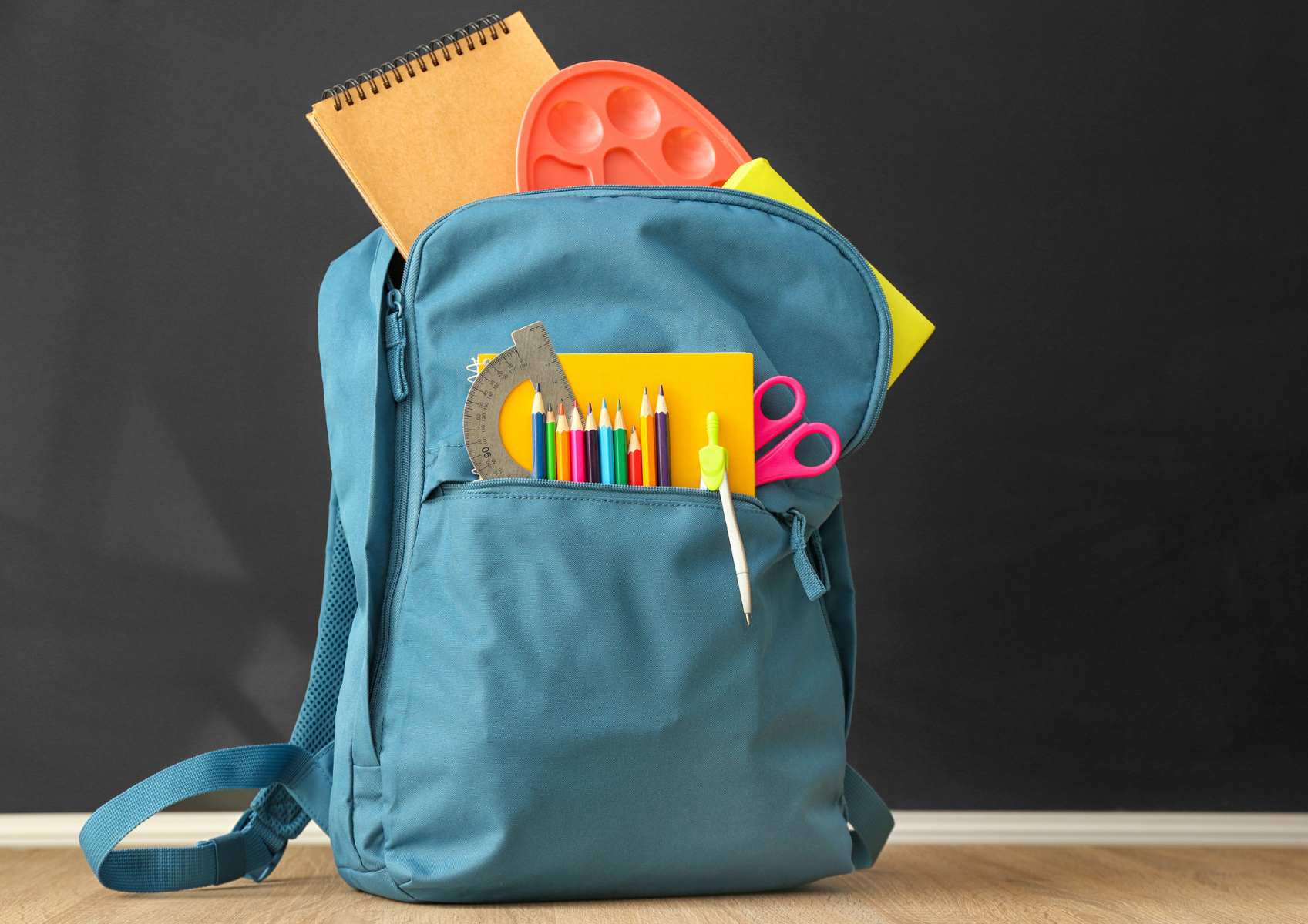 Na czarnym "tablicowm" tle znajduje się niebieski plecak z którego wystają przybory plastyczne, długopisy, kartki, ekierka, nożyczki itp.
