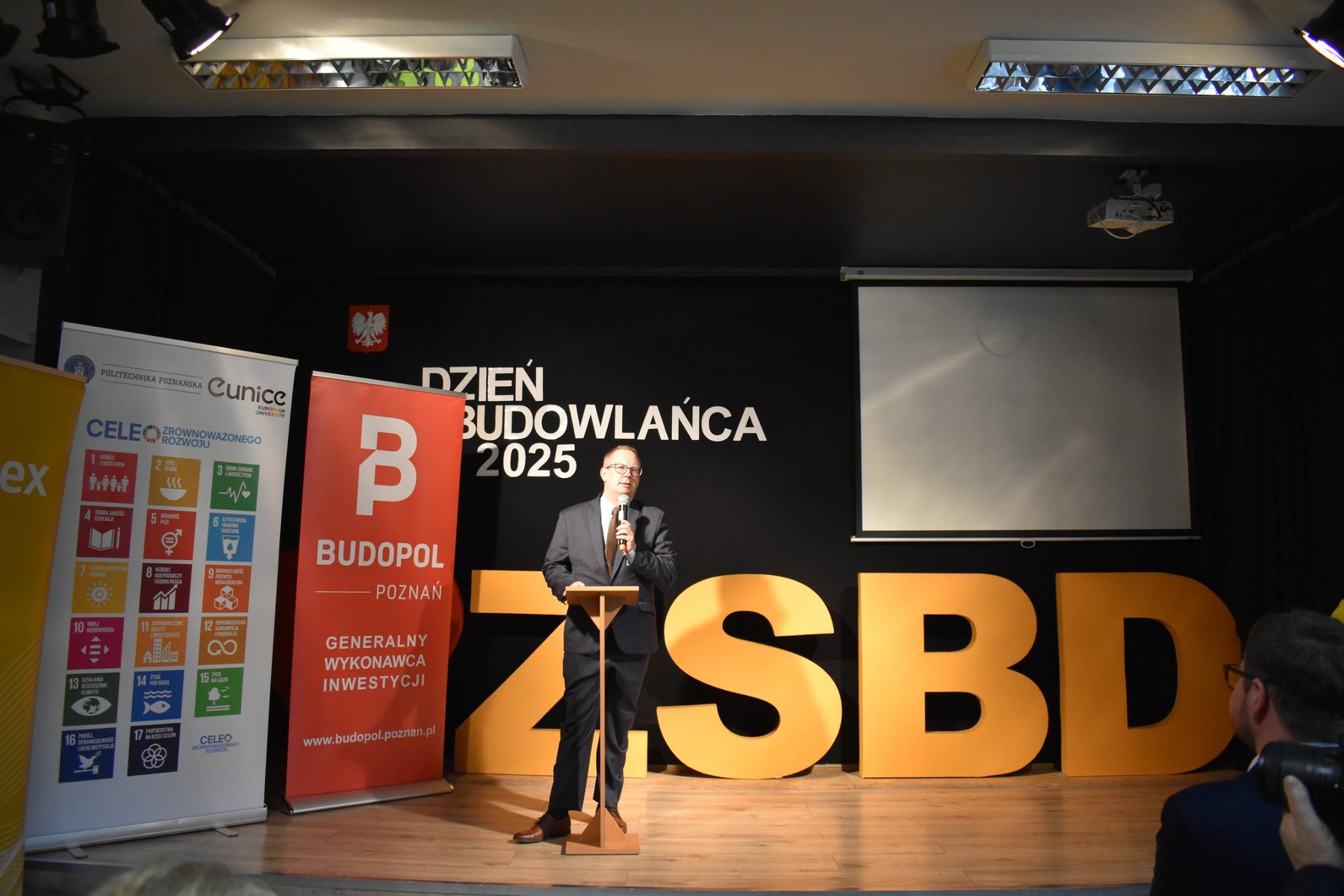 Na czarnym tle sceny przemawia Dyrektor Zespołu Szkół Budowlano-Drzewnych