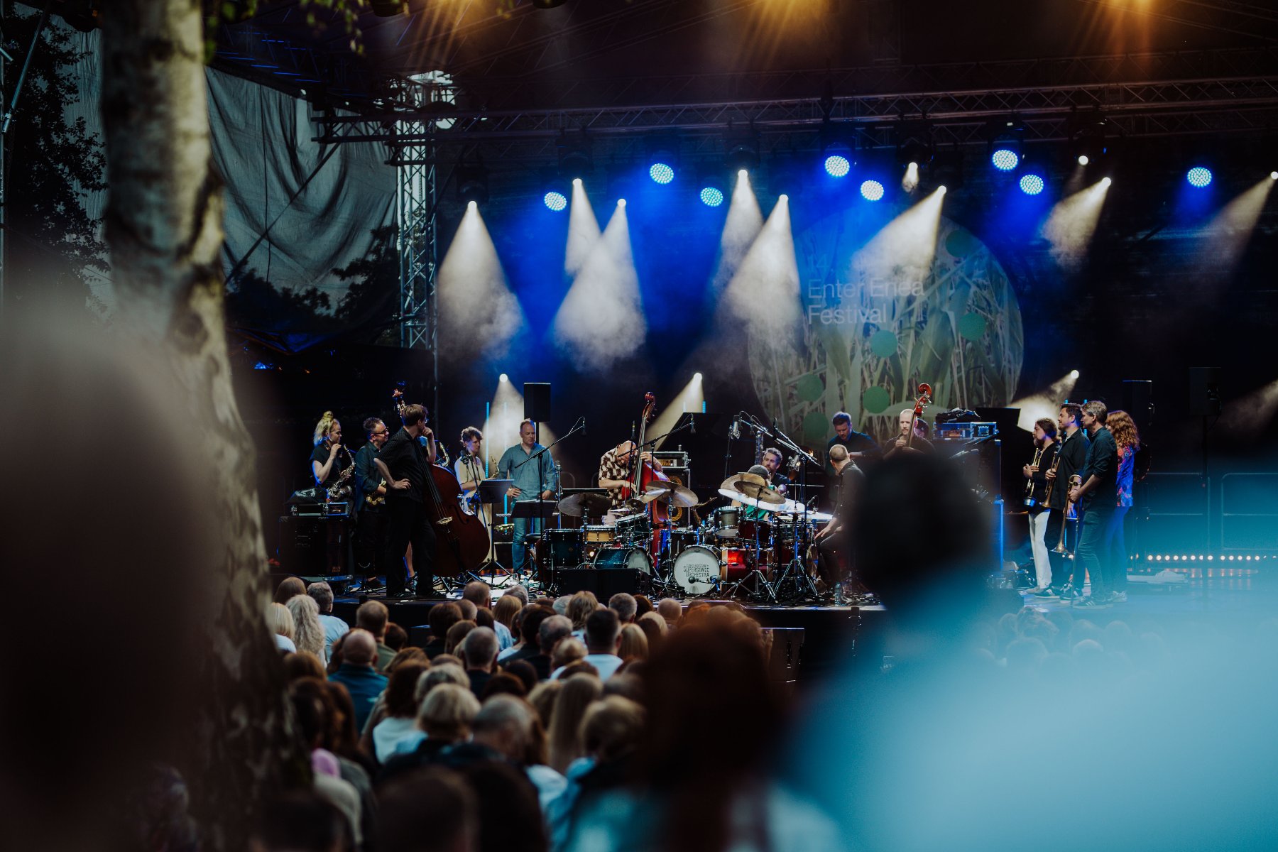 W przyszłym tygodniu rzusza kolejny festiwal muzyczny. Od 15 do 18 czerwca wybrzeża Jeziora Strzeszyńskiego rozgrzeje Enter Enea Festival. Na pierwszym planie głowy słuchaczy, którzy siedzą pod sceną i oglądają koncert. Na scenie zespół składający się z wielu muzyków. Scena jest oświetlona bialymi i niebieskimi światłami.