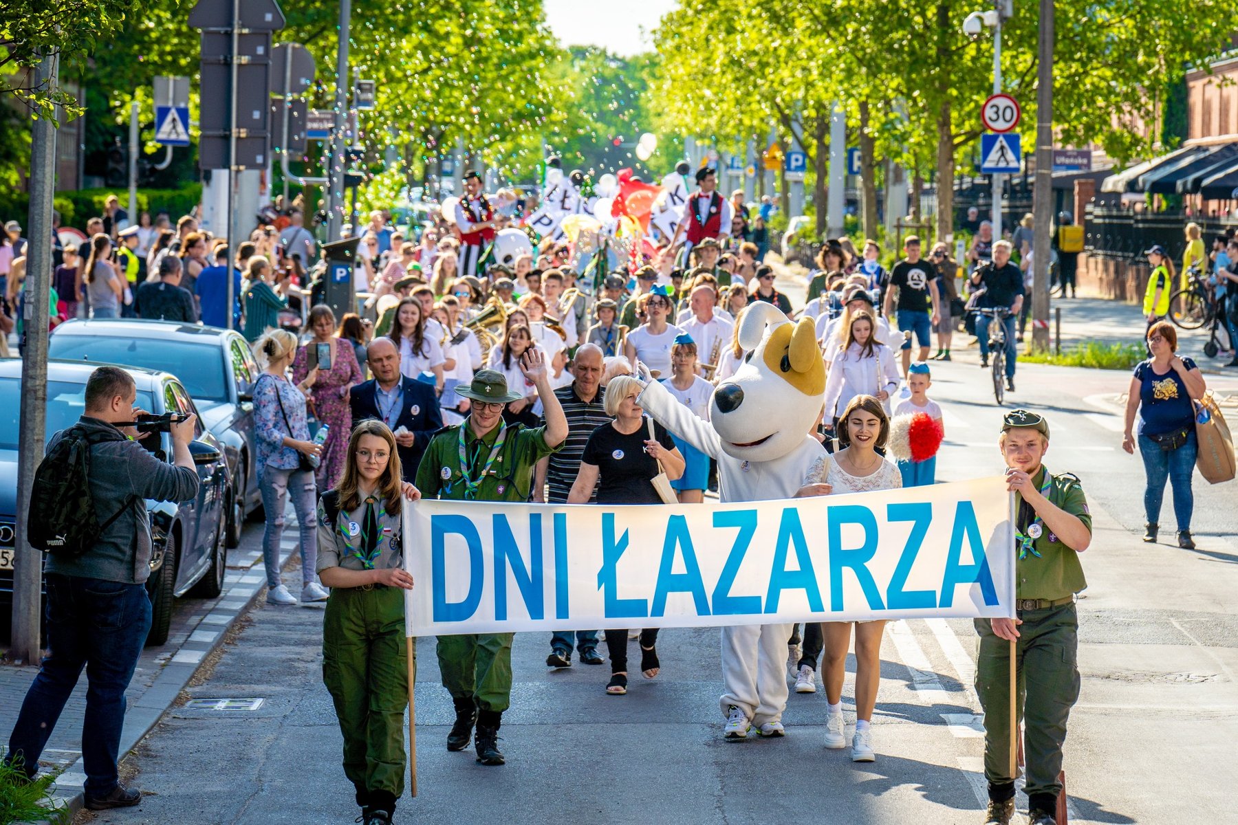 Dzisiaj o 17:00 uroczysty korowód przejdzie główną ulicą Łazarza i rozpocznie oficjalne świętowanie i letni sezon na Łazarzu. Na pierwszym planie idzie grupa harcerzy i trzyma baner "Dni Łazarza". Za nimi idzie reszta korowodu, która składa się z mieszkańców Poznania. Po drodze stoją inni ludzie, którzy przyglądają się wydarzeniu.