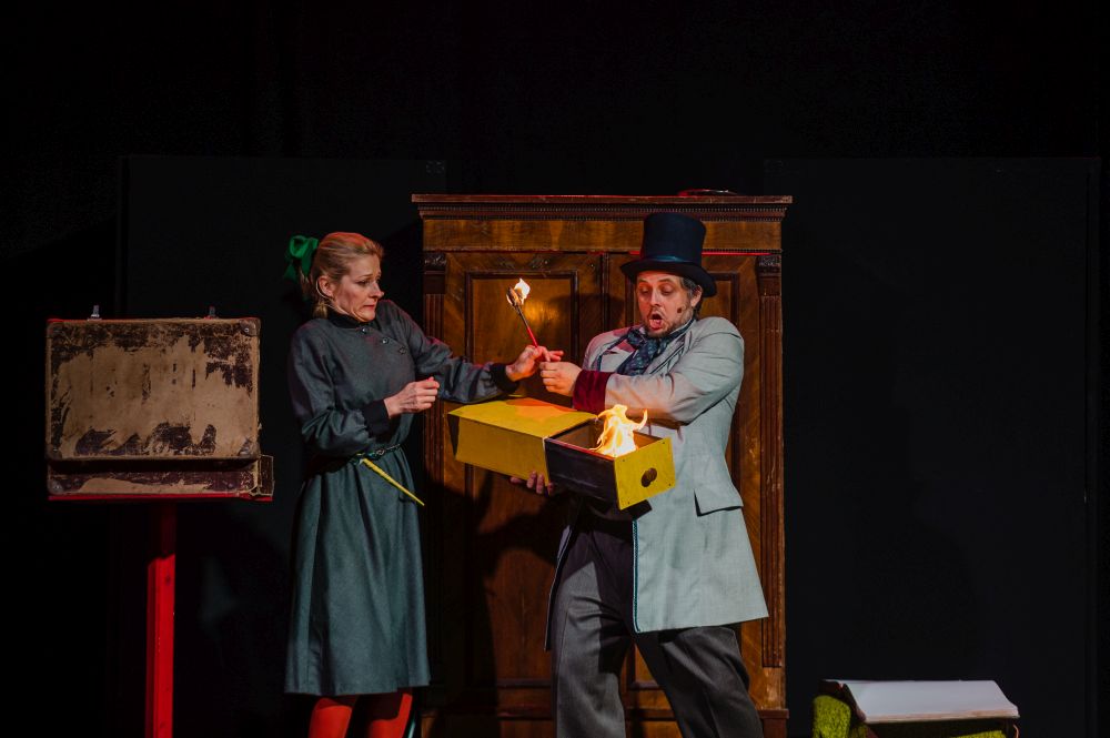 Na scenie kobieta i mężczyzna. Podają sobie dużą zapaloną zapałkę, mężczyna trzyma duże otwarte pudełko od zapałek, w którym widać ogień.