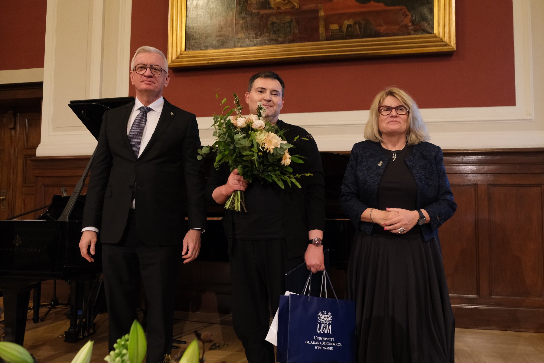 Poznańska Nagroda Literacka, foto: Przemysław Stanula na tle auli stoi dwóch mężczyzn i kobieta. Mężczyzna stojący pośrodku trzyma w jednej ręce bukiet kwiatów a w drugiej torbę z upominkiem