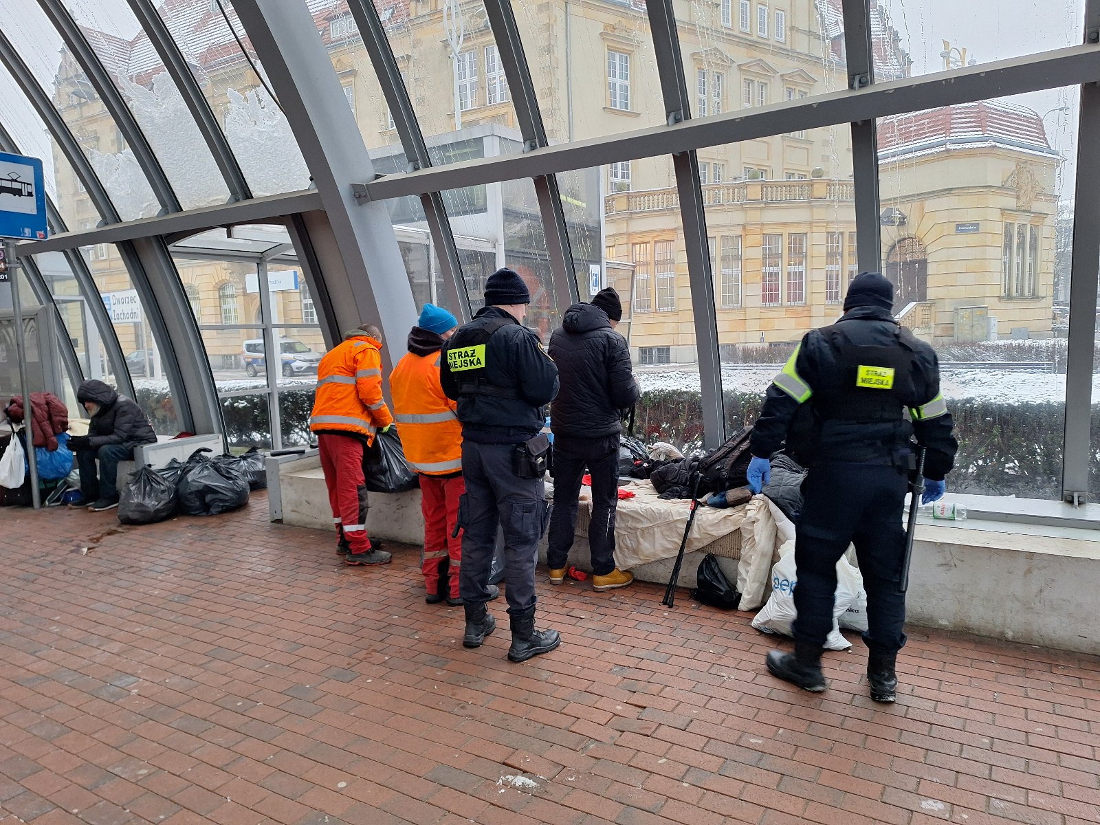 Na zdjęciu dwóch strażników miejskich, dwóch pracowników pogotowia czystości w pomarańczowych uniformach i dwóch mężczyzn w kryzysie bezdomności na dworcu zachodnim. Przy nich worki ze śmieciami i leżące ubrania. W tle przez szyby dworca widać budynek poczty polskiej.