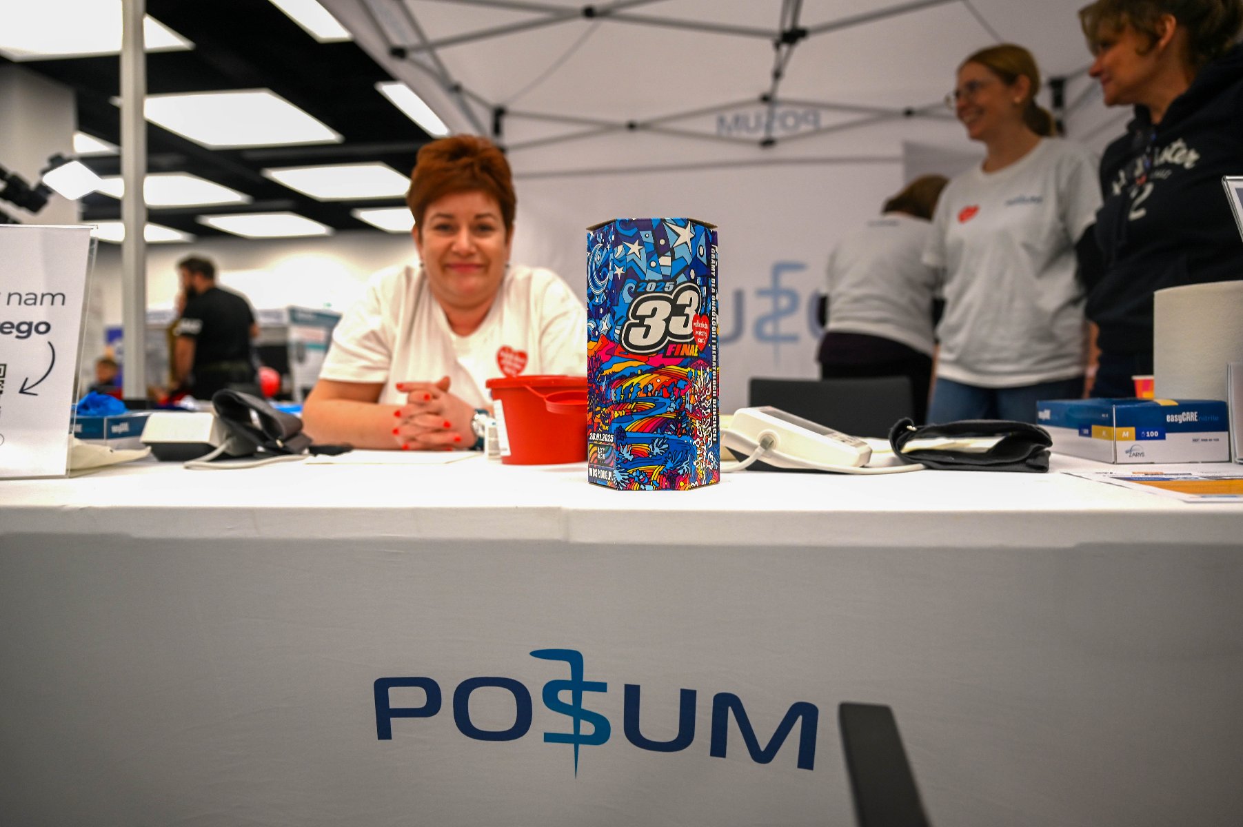 Na zdjęciu kobieta w medycznym fartuchu przytyka coś do twarzy kobiecie siedzącej tyłem do obiektywu