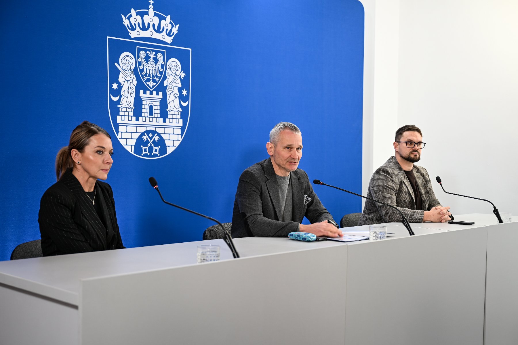 Na zdjęciu mężczyzna i kobieta za stołem konferencyjnym, za nimi ścianka z herbem miasta oraz grafika przedstawiająca dziecko