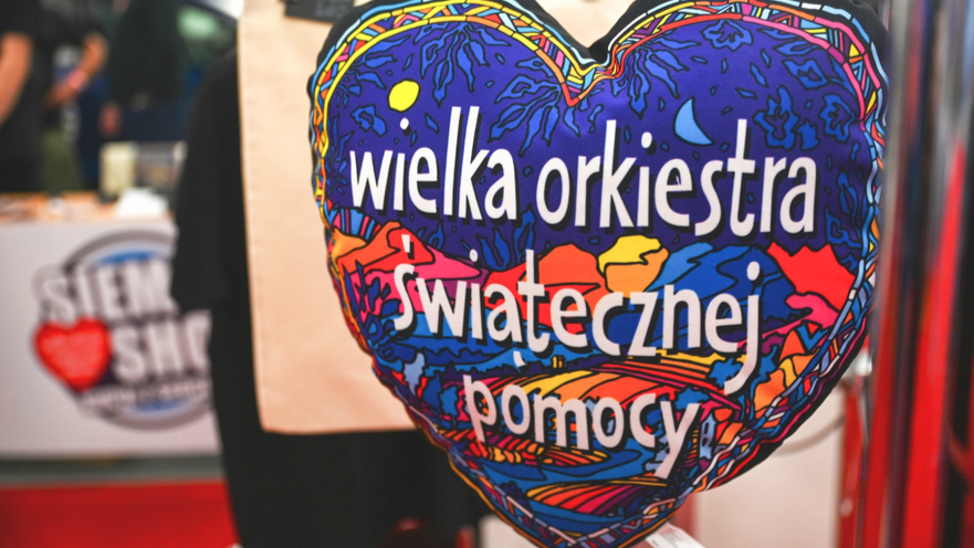 Na zdjęciu poduszka w kształcie kolorowego serca z napisem: "Wielka Orkiestra Świątecznej Pomocy". Tło rozmazane.