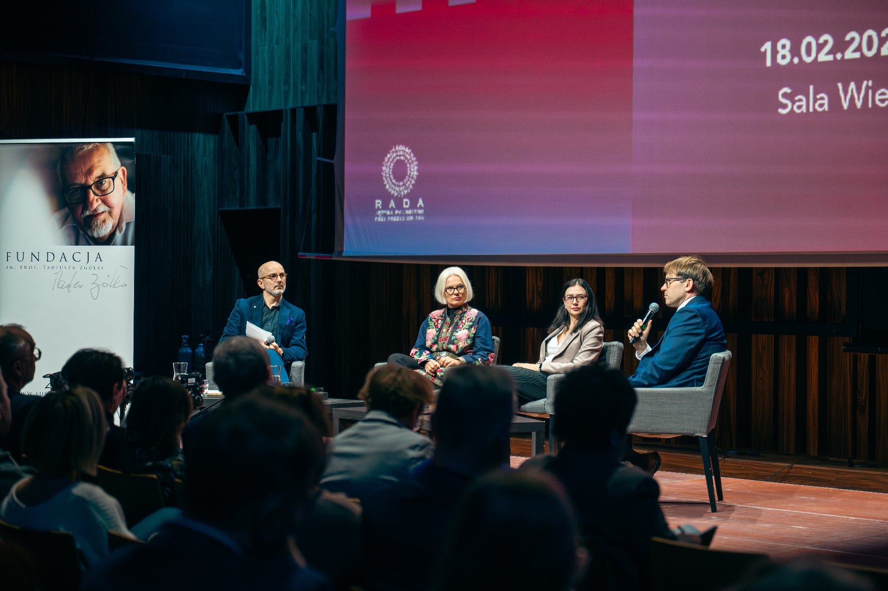 Na zdjęciu sala wypełniona publicznością, przy mównicy mężczyzna, za nim duzy ekran