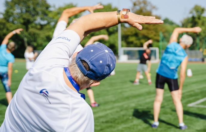 Na zdjęciu seniorzy uprawiający gimnastykę na świeżym powietrzu, stojący tyłem do obiektywu