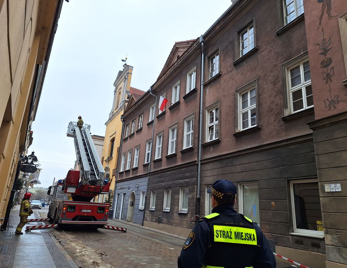 na zdjęciu strażnik miejski tyłem do obiektywu obserwuje strażaków i wóz strażacki z drabiną oraz wysięgnikiem koszowym na ul. Żydowskiej