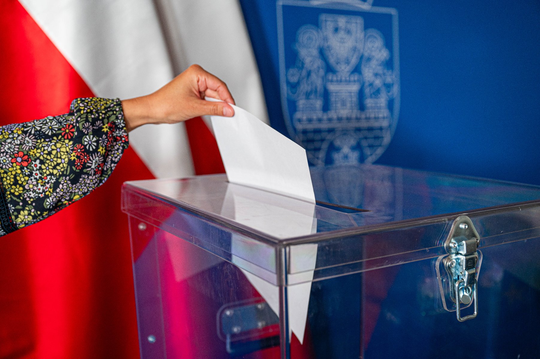 Wybory 2023 - upływa ważny termin dla studentów Na zdjęciu widać urnę wyborczą oraz rękę, która wrzuca do urny kartę do głosowania. W tle herb Poznania oraz flagi Polski.