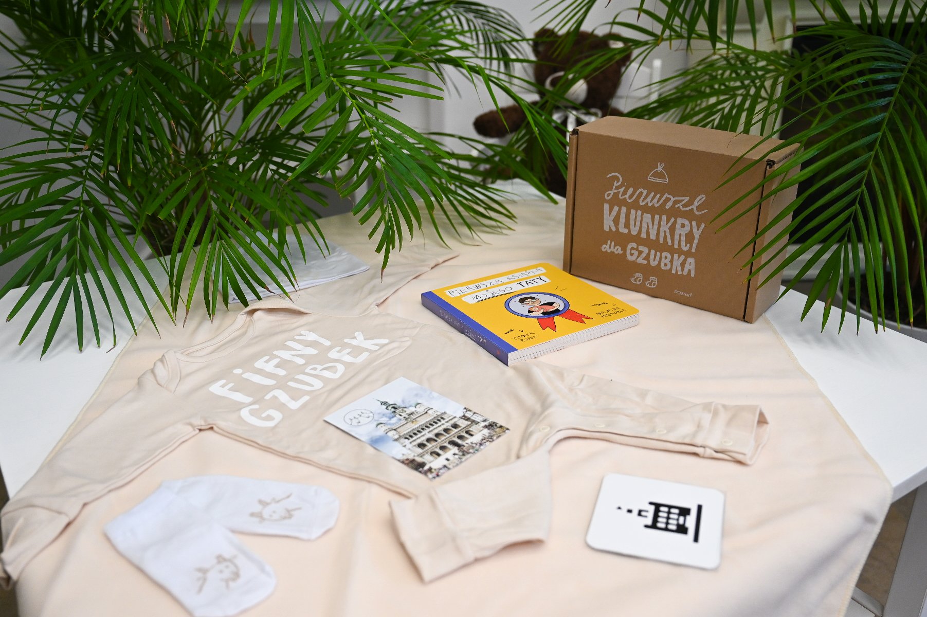 Na zdjęciu wyprawka: na stoliku rozłożone śpioszki i inne elementy wyprawki, w tle widać rośliny