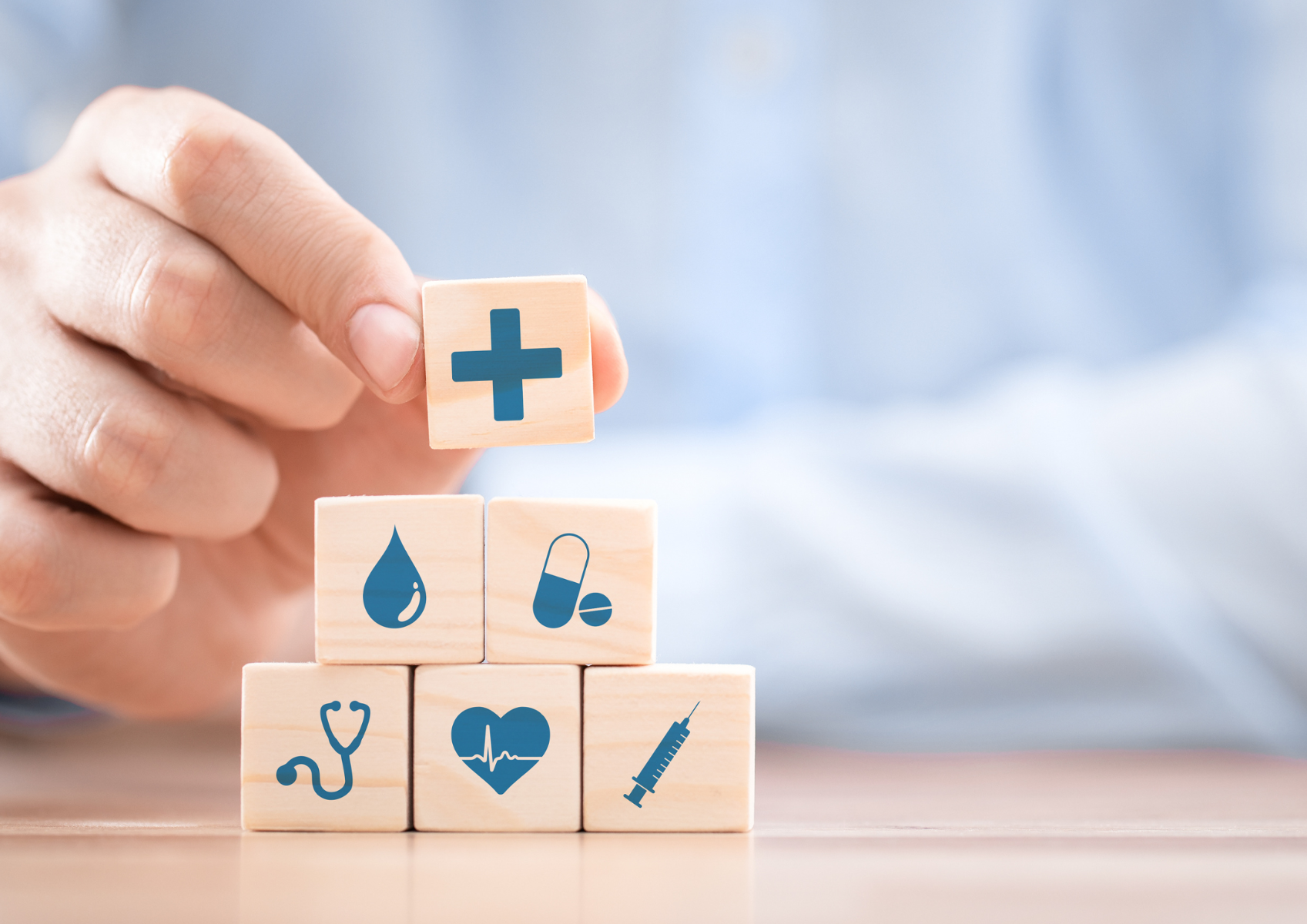 Na zdjęciu znajduje się ręka ustawiająca drewniane klocki, na których umieszczone są elementy kojarzące się ze zdrowiem, tj. stetoskop, krzyż, kropla