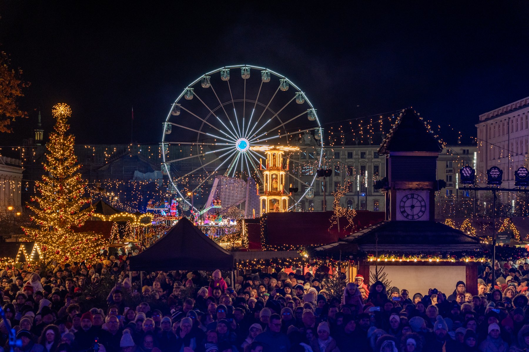 Nocny widok na zatłoczony Jarmark Świąteczny z dużą, jasno oświetloną choinką po lewej i diabelskim młynem w centrum kadru. W dolnej części zdjęcia widoczny jest duży tłum ludzi, a w tle udekorowane światłami budynki.