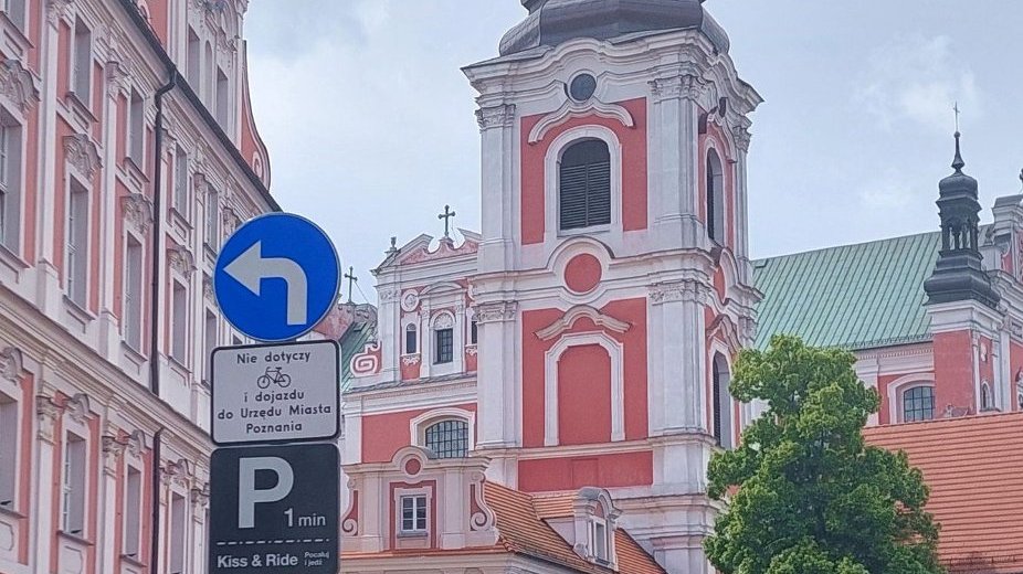 Na zdjęciu jest znak drogowy nakaz skrętu w lewo oraz tabliczka kiss and ride postój do 1 minuty, w tle budynek poznańskiego urzedu miasta z różowo-białą elewacją oraz drzewo