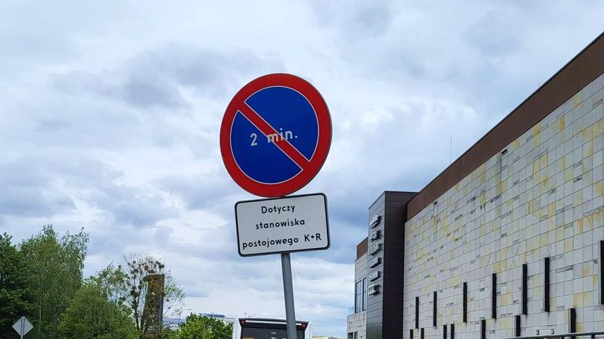 Na zdjęciu jest znak drogowy B-35 zakaz postoju powyżej dwóch minut, w tle niebo oraz fragment budynku galerii handlowej Posnania oraz drzewa