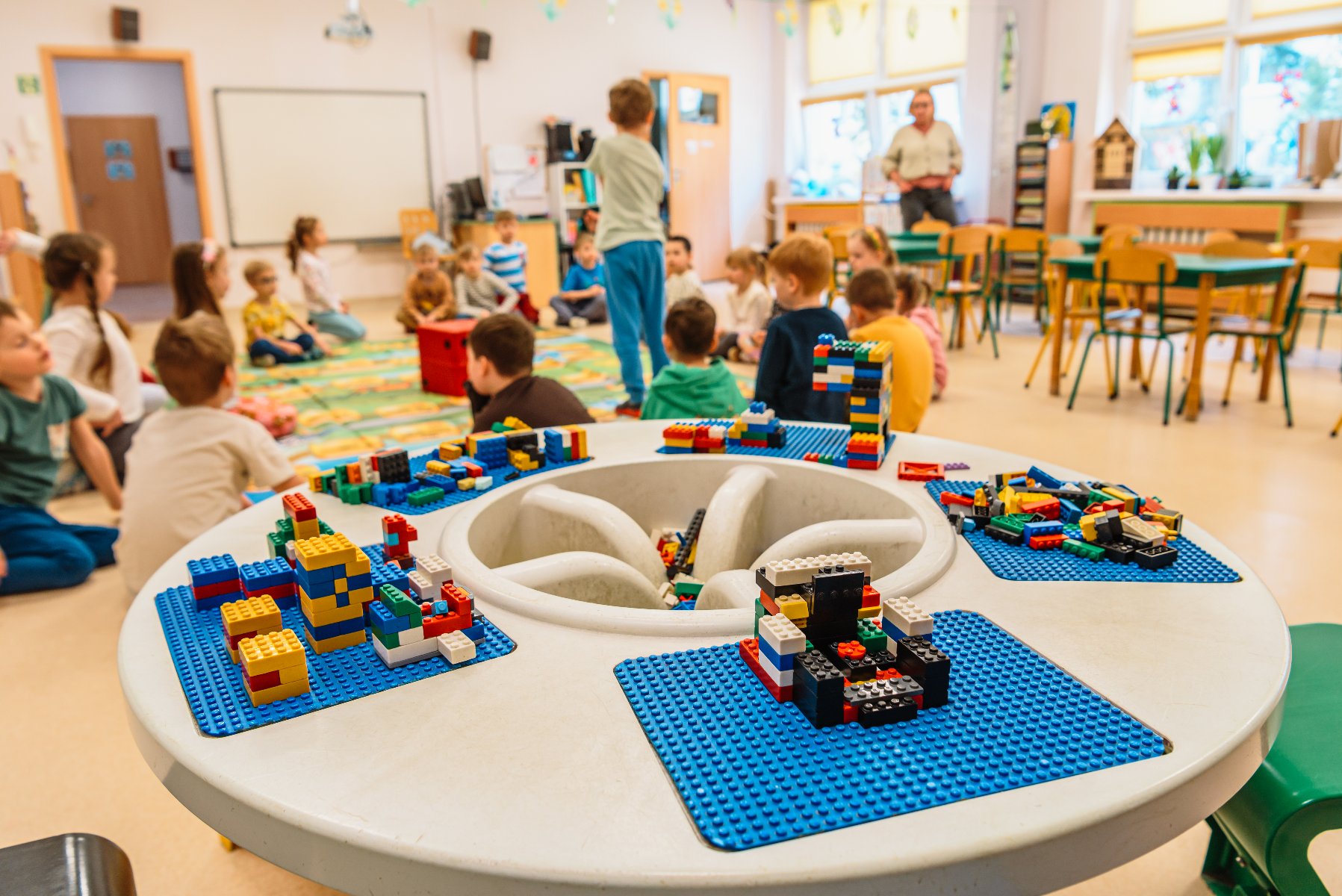 Zdjęcie przedstawia dzieci w sali przedszkolnej siedzące na dywanie. Na pierwszym planie znajduje się stół, na którym leżą klocki lego. - grafika artykułu