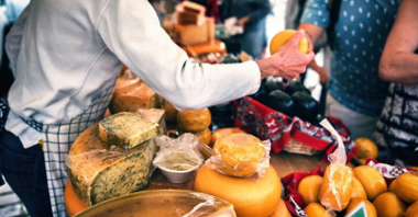 Zdjęcie przedstawia stoisko z serami oraz dłoń podającą jeden z produktów.
