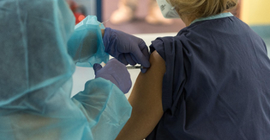 Na zdjęciu szczepienie: osoba w medycznym stroju szczepi kobietę; nie widać ich twarzy, w centrum zdjęcia dłonie ze szczepionką