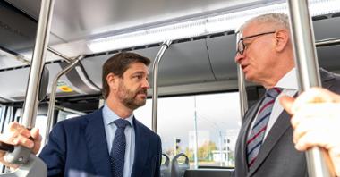 Na zdjęciu dwóch mężczyzn w autobusie, rozmawiają, trzymając się poręczy