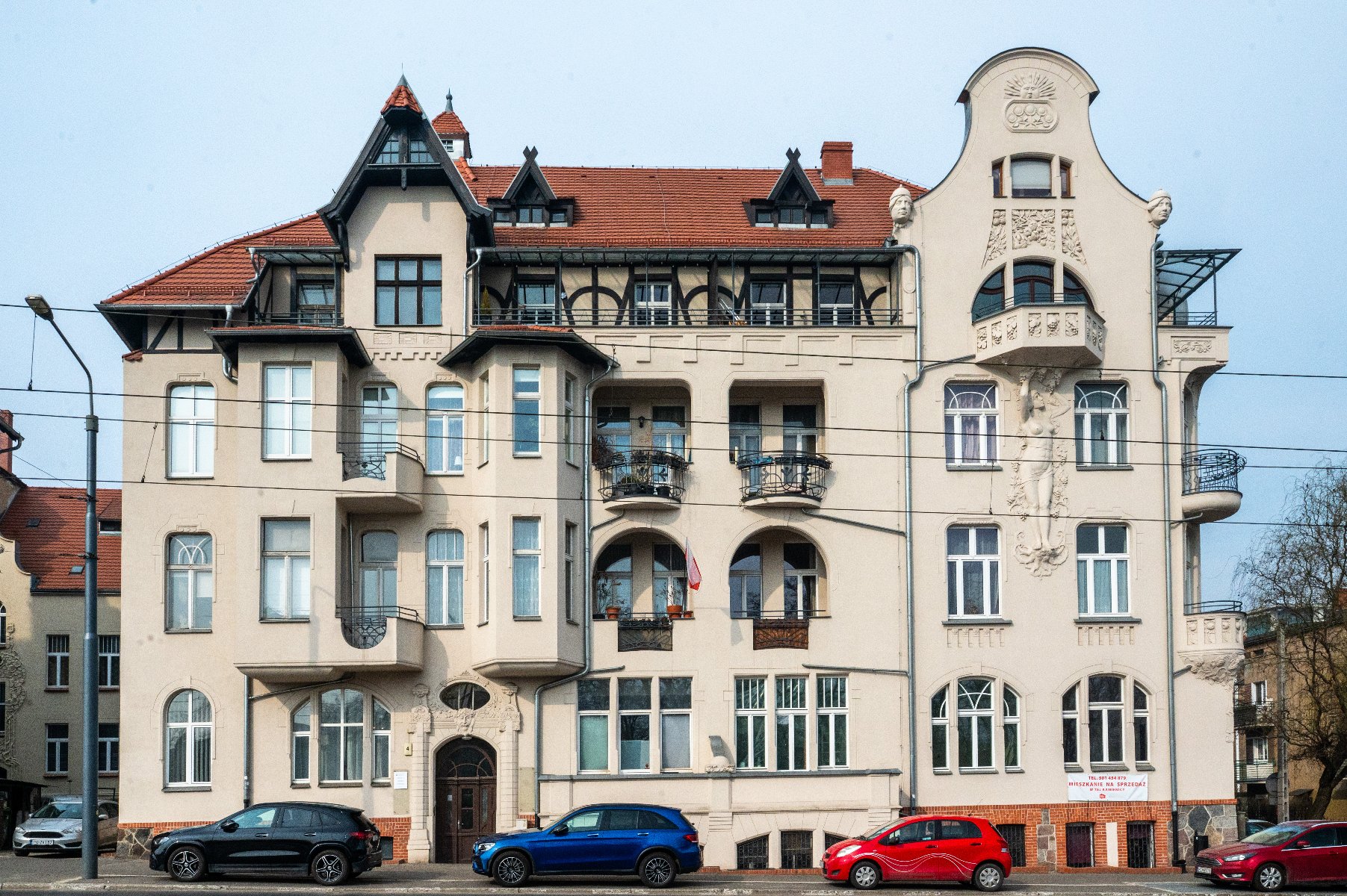 Wielopiętrowy budynek mieszkalny o bogato zdobionej architekturze, łączący różne style, w tym secesję. Na elewacji znajdują się dekoracyjne detale, takie jak misternie rzeźbienia, balkony z kutymi żelaznymi balustradami oraz różnorodne kształty i rozmiary okien. Dach jest stromo spadzisty z lukarnami i szczytami. Przed budynkiem zaparkowanych jest kilka samochodów, widoczne są latarnia uliczna i linie energetyczne. - grafika artykułu