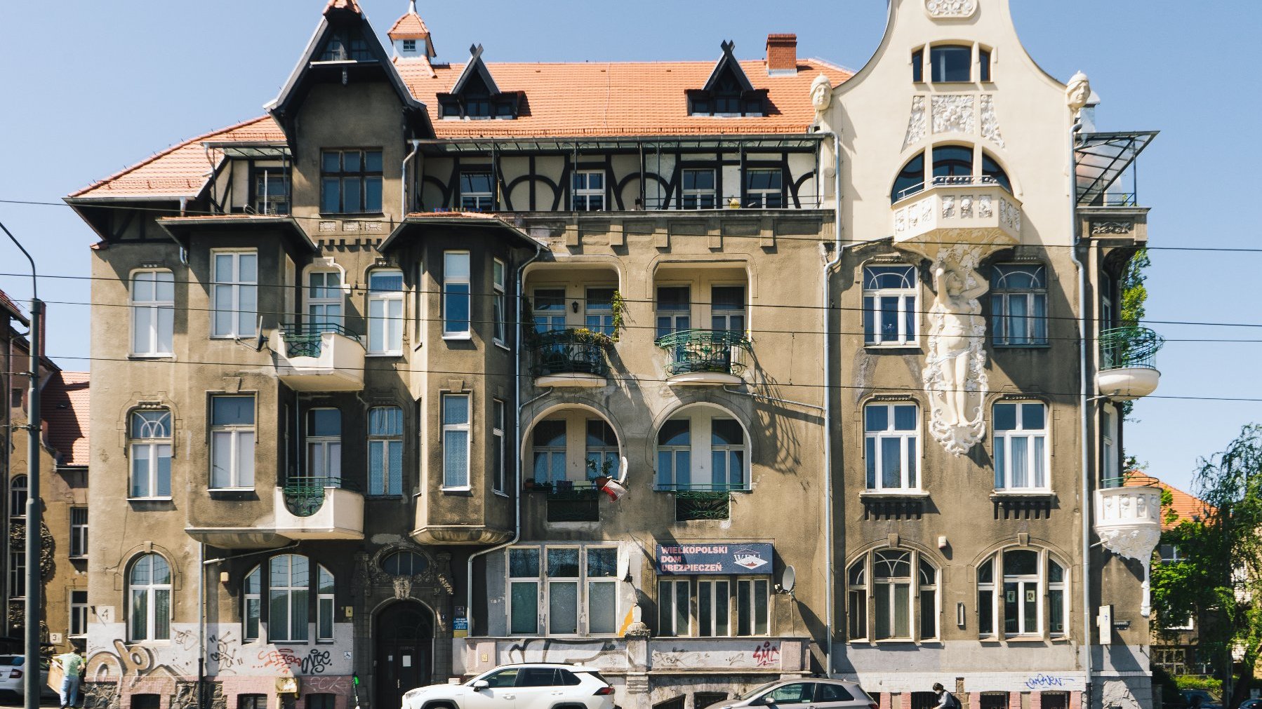 Wielopiętrowy budynek mieszkalny o bogato zdobionej architekturze, łączący różne style, w tym secesję. Na elewacji znajdują się dekoracyjne detale, takie jak misternie rzeźbienia, balkony z kutymi żelaznymi balustradami oraz różnorodne kształty i rozmiary okien. Dach jest stromo spadzisty z lukarnami i szczytami. Przed budynkiem zaparkowane są dwa samochody, widoczne są również graffiti na dolnej części elewacji.