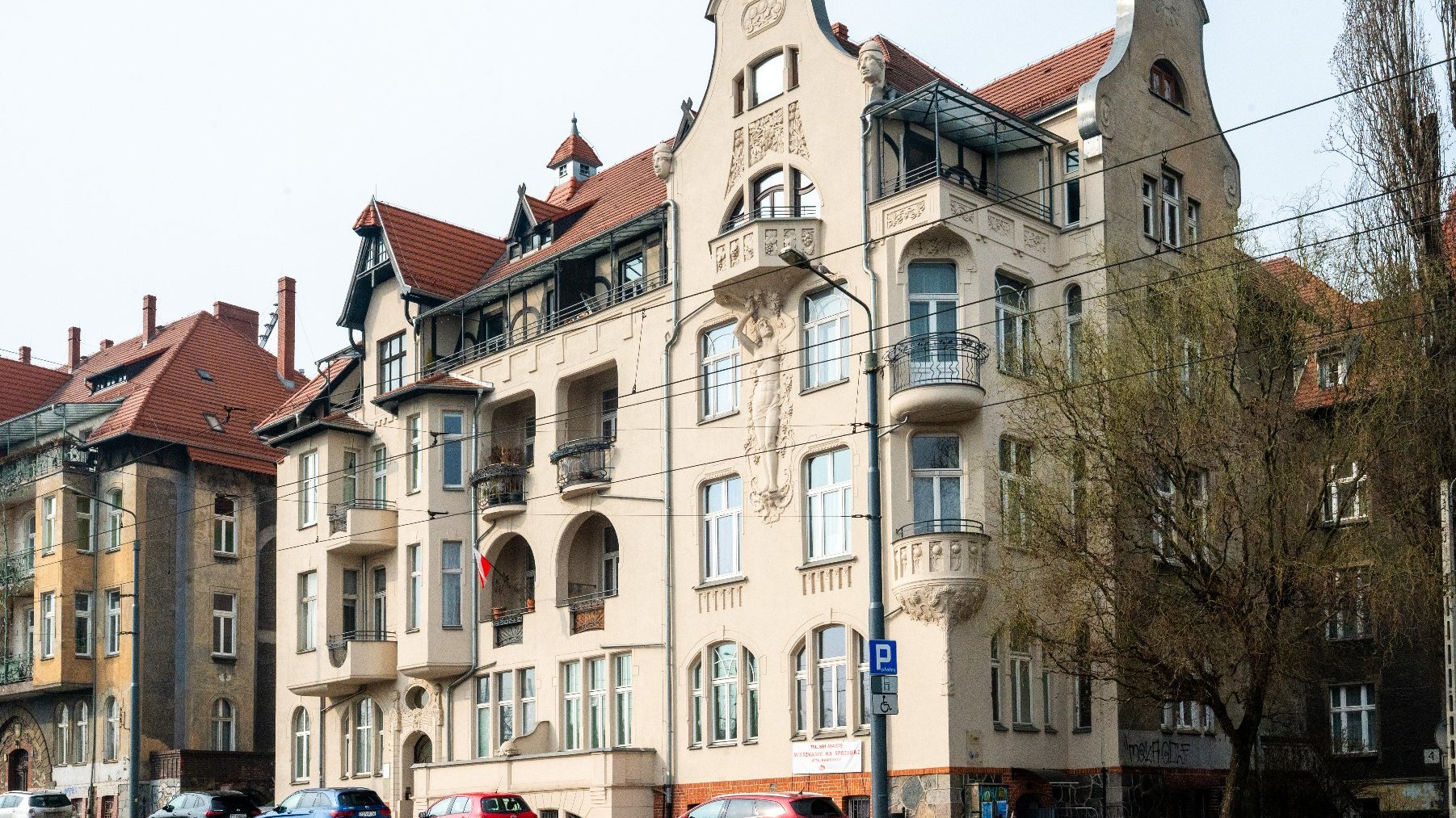 Widok ulicy z budynkiem mieszkalnym o bogatej, ozdobnej architekturze. Budynek ma beżową elewację z misternymi detalami, w tym dekoracyjnymi rzeźbami i balkonami. Dach jest czerwony, z wieloma szczytami i oknami dachowymi. Przed budynkiem zaparkowanych jest kilka samochodów, widoczny jest również rower. Budynek jest otoczony innymi budynkami mieszkalnymi o podobnym stylu architektonicznym. Na zdjęciu widać niebieski znak parkingowy z symbolem niepełnosprawności, oznaczający dostępne miejsce parkingowe.