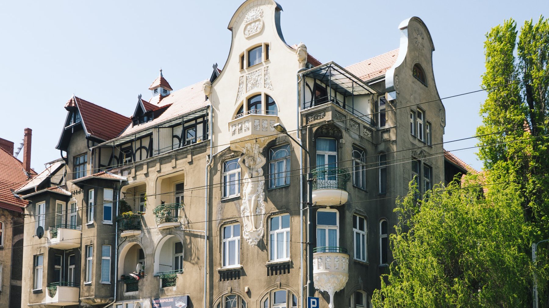 Wielopiętrowy budynek o eklektycznym stylu architektonicznym. Fasada ozdobiona jest różnymi elementami dekoracyjnymi, takimi jak łukowe okna, balkony z misternymi balustradami oraz rzeźbione reliefy. Dach pokryty jest czerwonymi dachówkami, a na nim widoczne są liczne lukarny. Budynek znajduje się w miejskim otoczeniu, na pierwszym planie widać drzewa i linie energetyczne.