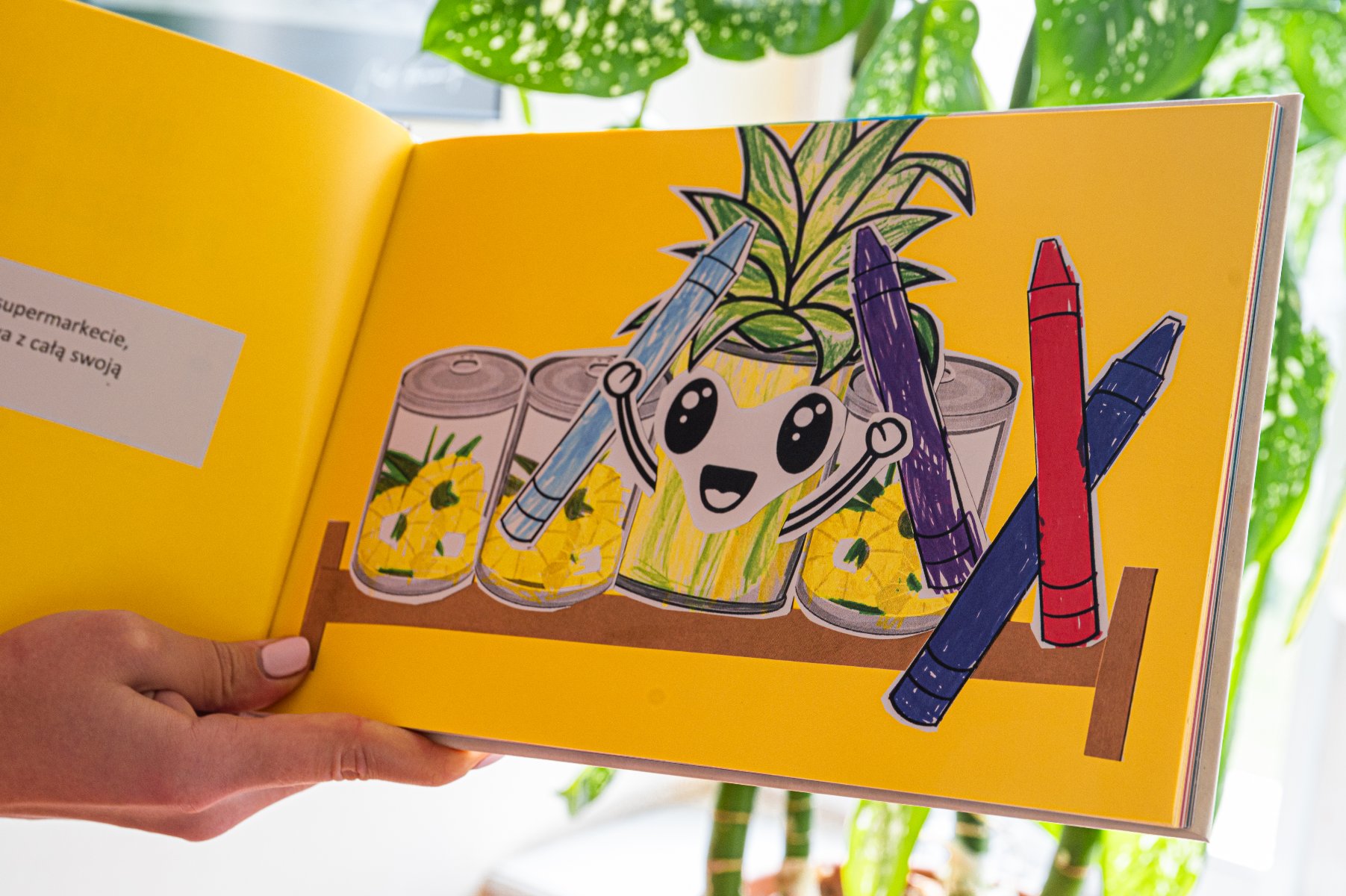 Książka dla dzieci z jasnym żółtym tłem. Na prawej stronie ilustracja uśmiechniętego ananas z dużymi, wyrazistymi oczami i szerokim uśmiechem. Ananas trzyma dwa kredki: niebieską i fioletową, a obok, na półce, opierają się dwie inne kredki: czerwona i niebieska. Na półce znajdują się również trzy puszki z plasterkami ananasa. Na lewej stronie jest mały blok tekstu w języku polskim: "supermarkecie, z całą swoją". Książka jest trzymana przez osobę z różowym lakierem do paznokci. - grafika artykułu