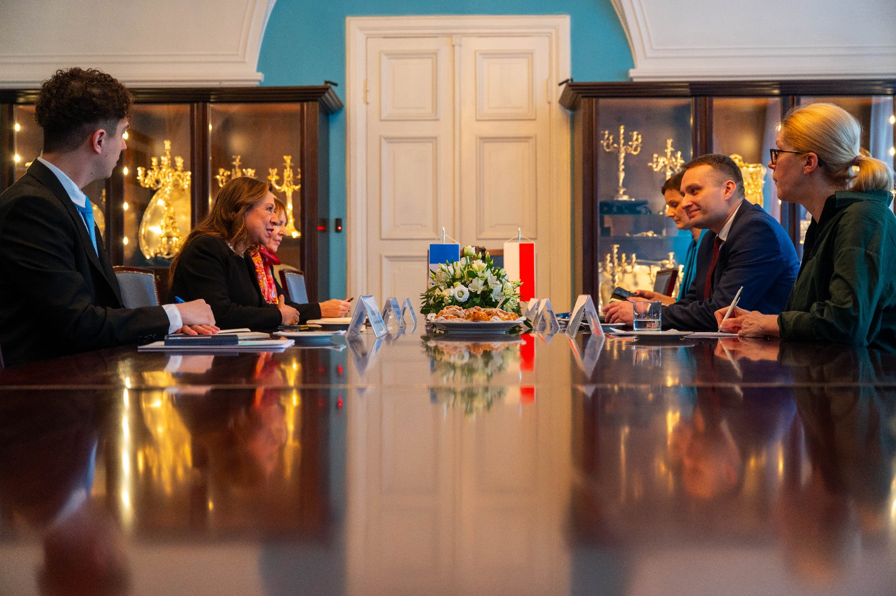 Formalne spotkanie odbywające się w elegancko udekorowanym pomieszczeniu. Przy dużym drewnianym stole siedzą cztery osoby, po dwie z każdej strony, twarzą do siebie. Na stole widoczne są tabliczki z nazwiskami i dokumenty, co sugeruje, że jest to spotkanie dyplomatyczne lub biznesowe. Na środku stołu znajduje się kompozycja kwiatowa oraz dwie małe flagi - grafika artykułu