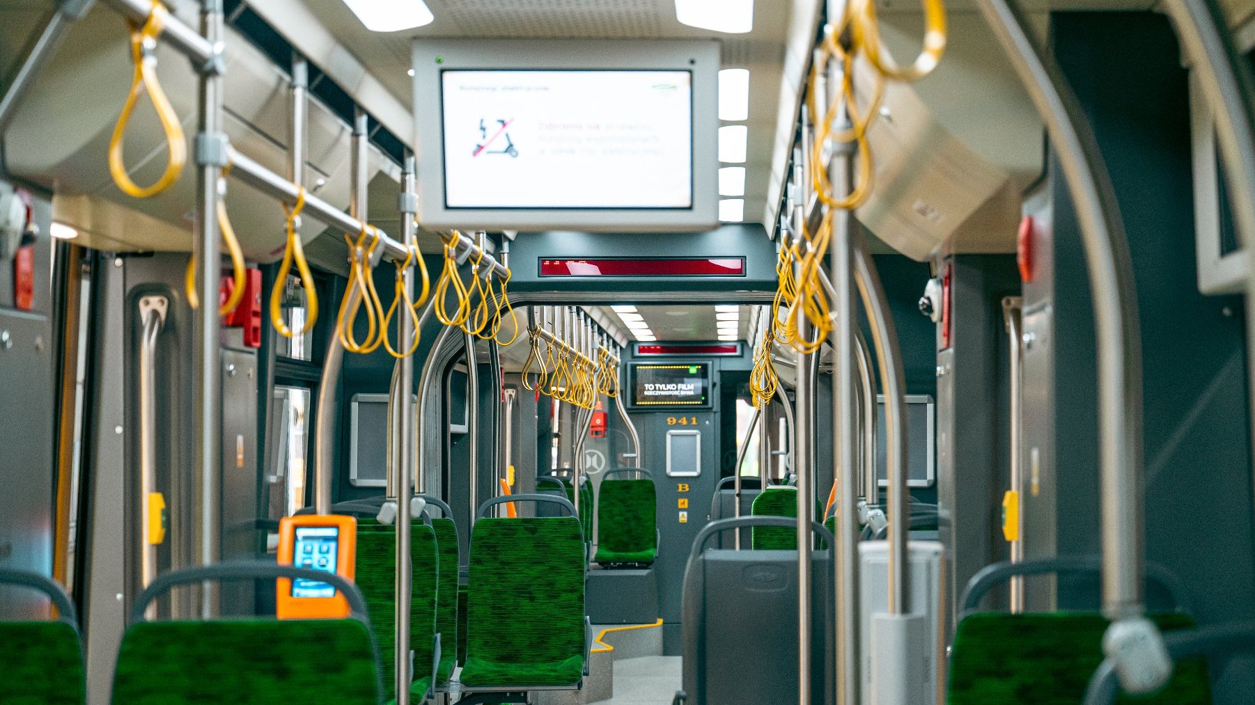 Na zdjęciu wnętrze tramwaju, widać siedzenia, poręcze i monitor wiszący pod sufitem