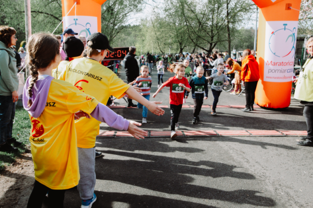 Dzieci uczestniczą w biegu, przekraczając linię mety oznaczoną pomarańczowym, dmuchanym łukiem z napisem "Maratonica pomiar czasu" i adresem strony internetowej "www.maratonypolskie.pl". - grafika artykułu