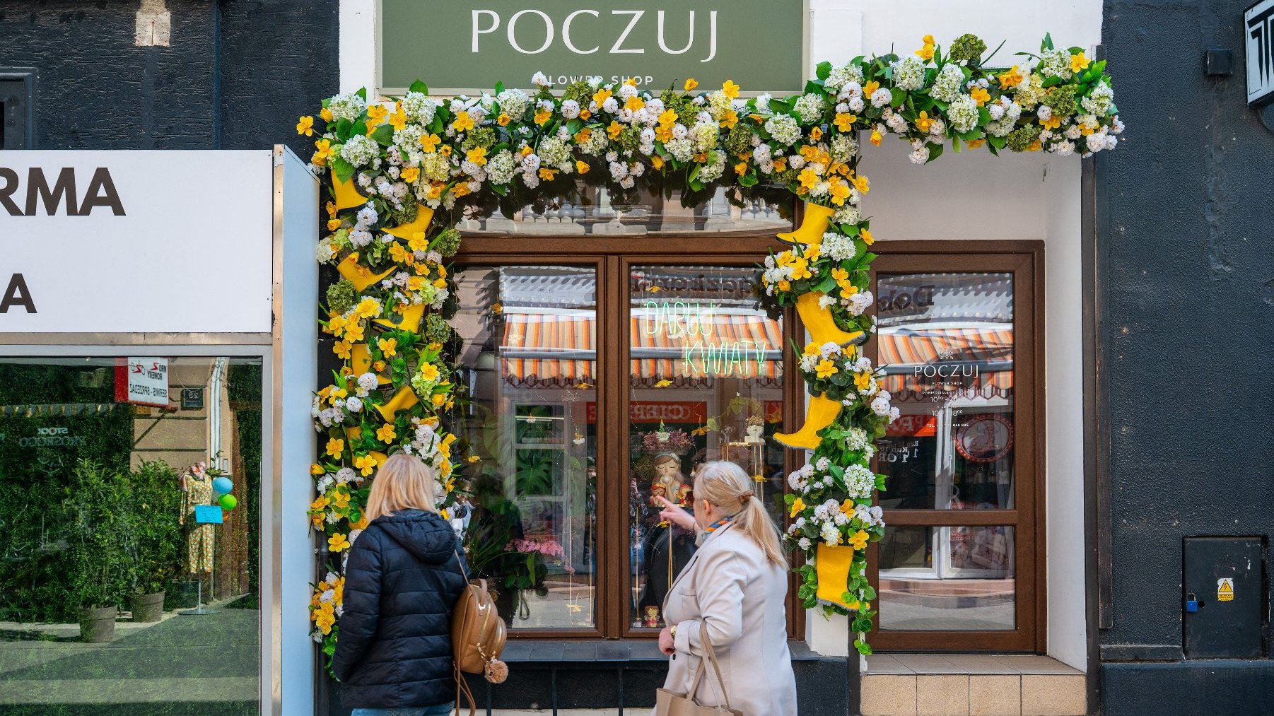Wejście do kwiaciarni o nazwie "POCZUJ". Nad drzwiami widnieje szyld "POCZUJ FLOWER SHOP". Przed wejściem znajduje się bogata aranżacja kwiatowa, głównie w odcieniach żółci i bieli, która zdobi fasadę i przyciąga wzrok przechodniów. Na wystawie sklepowej widoczny jest napis "DASZ KWIATY".