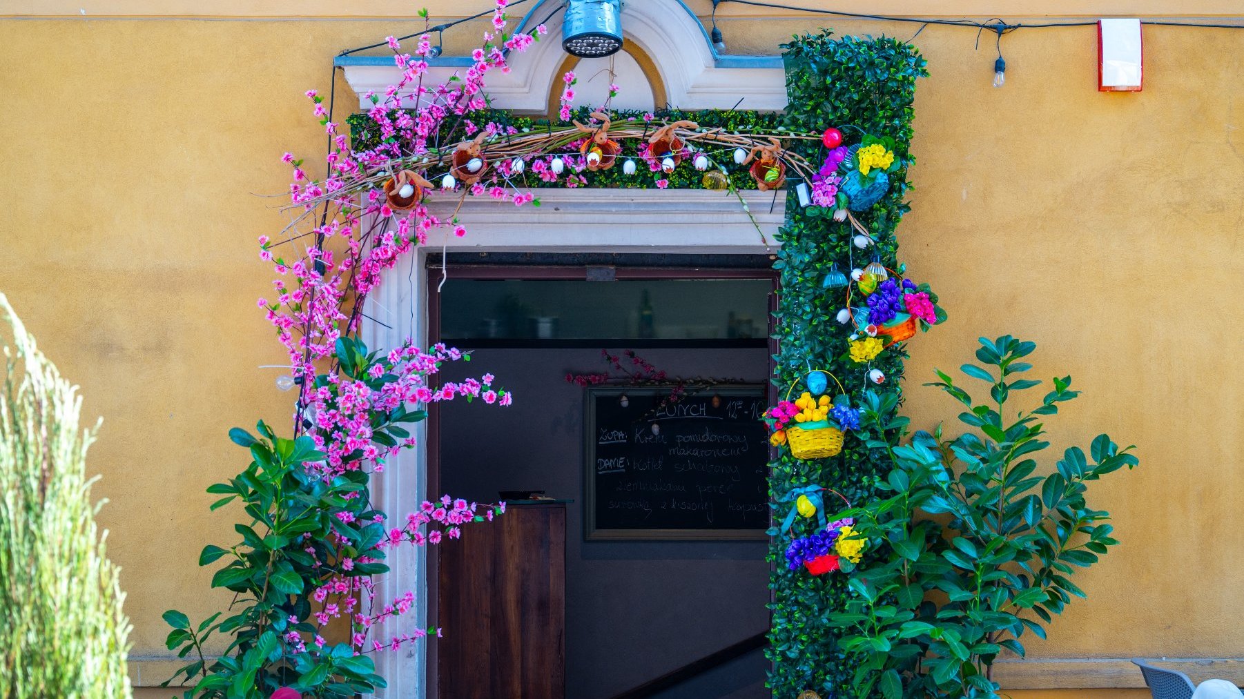 Wejście do budynku, prawdopodobnie restauracji lub kawiarni, ozdobione zielonymi liśćmi oraz kolorowymi kwiatami. Po lewej stronie drzwi widoczne są różowe kwiaty, a po prawej mieszanka żółtych, niebieskich i fioletowych kwiatów. Dodatkowe dekoracje, takie jak małe koszyki i ozdobne elementy, zwisają wokół wejścia.