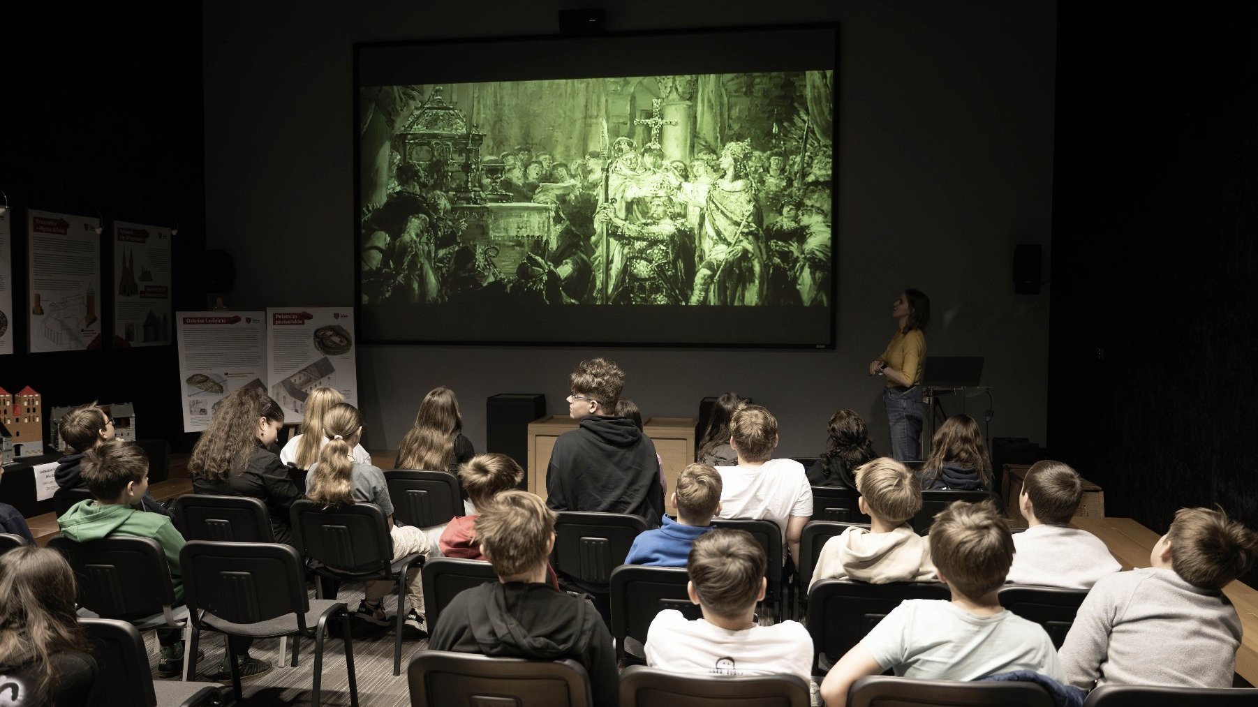 Pokój pełen dzieci oglądających prezentację na dużym ekranie. Na ekranie wyświetlana jest scena historyczna lub artystyczna, przedstawiona w zielonych odcieniach, z wieloma postaciami w stylu renesansowym lub średniowiecznym. Po prawej stronie ekranu stoi osoba prowadząca, która wyjaśnia lub omawia prezentowany materiał.