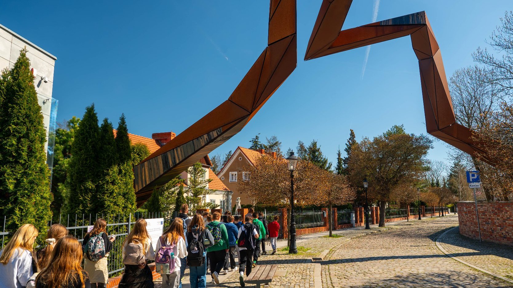 Ulica brukowana z charakterystycznym dużym metalowym rzeźbiarskim łukiem, który rozciąga się nad chodnikiem. Łuk ma geometryczny, nowoczesny design, nadający przestrzeni artystyczny akcent.