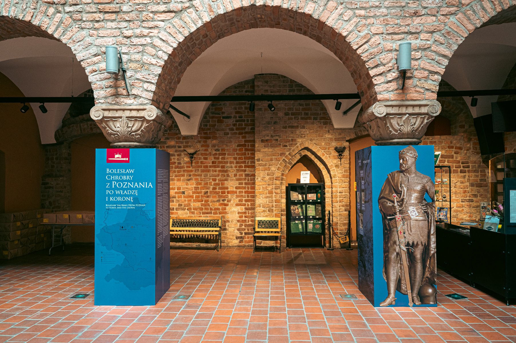 Zdjęcie przedstawia wnętrze muzeum. Widać na nim między innymi tablicę z informacją o wystawie oraz figurę króla. W tle znajdują się kasy i ławki. - grafika artykułu