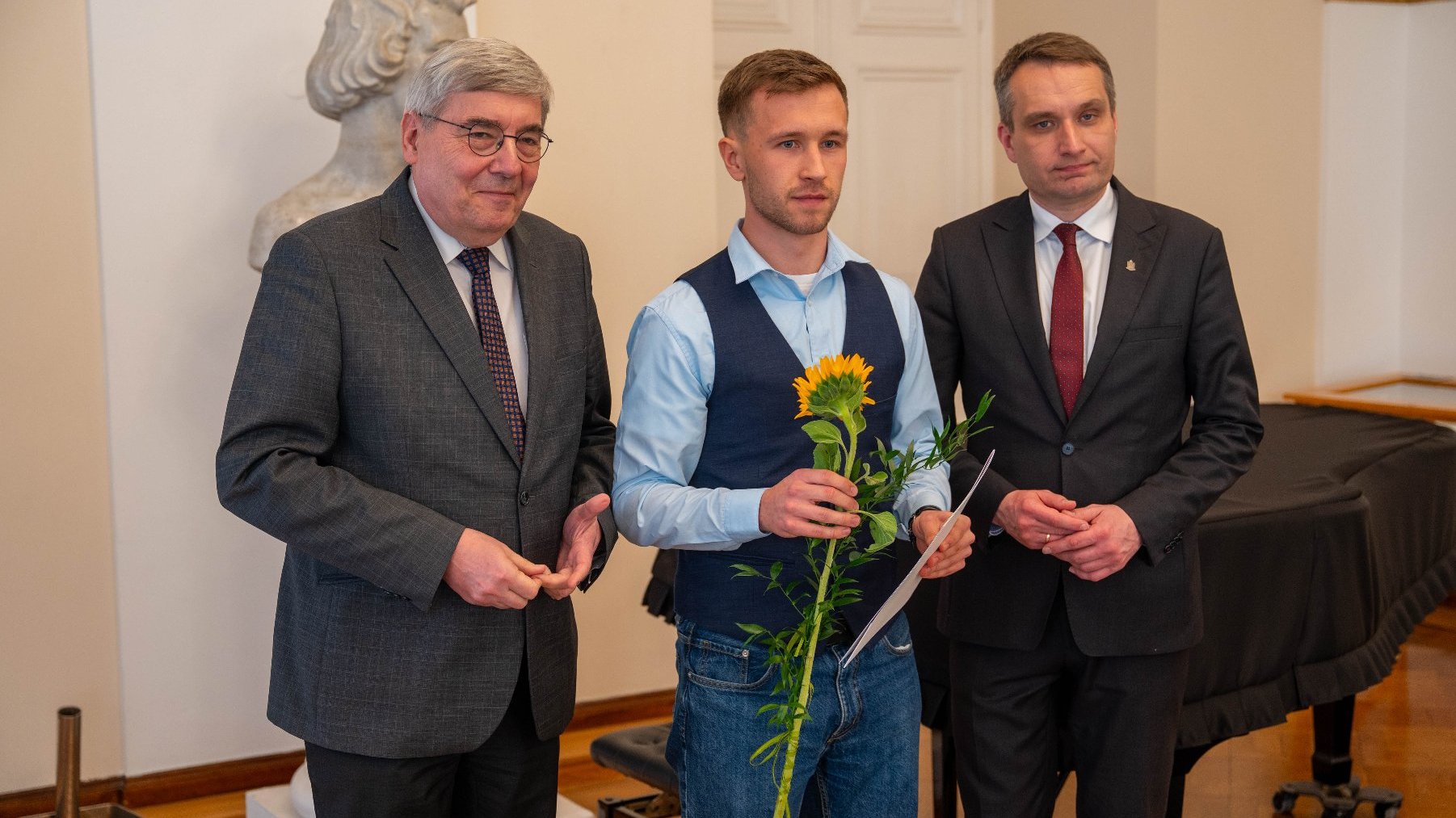 Trzech mężczyzn stoi w eleganckim wnętrzu. Osoba w środku trzyma słonecznik oraz kartkę papieru, podczas gdy pozostali są ubrani w garnitury i krawaty.